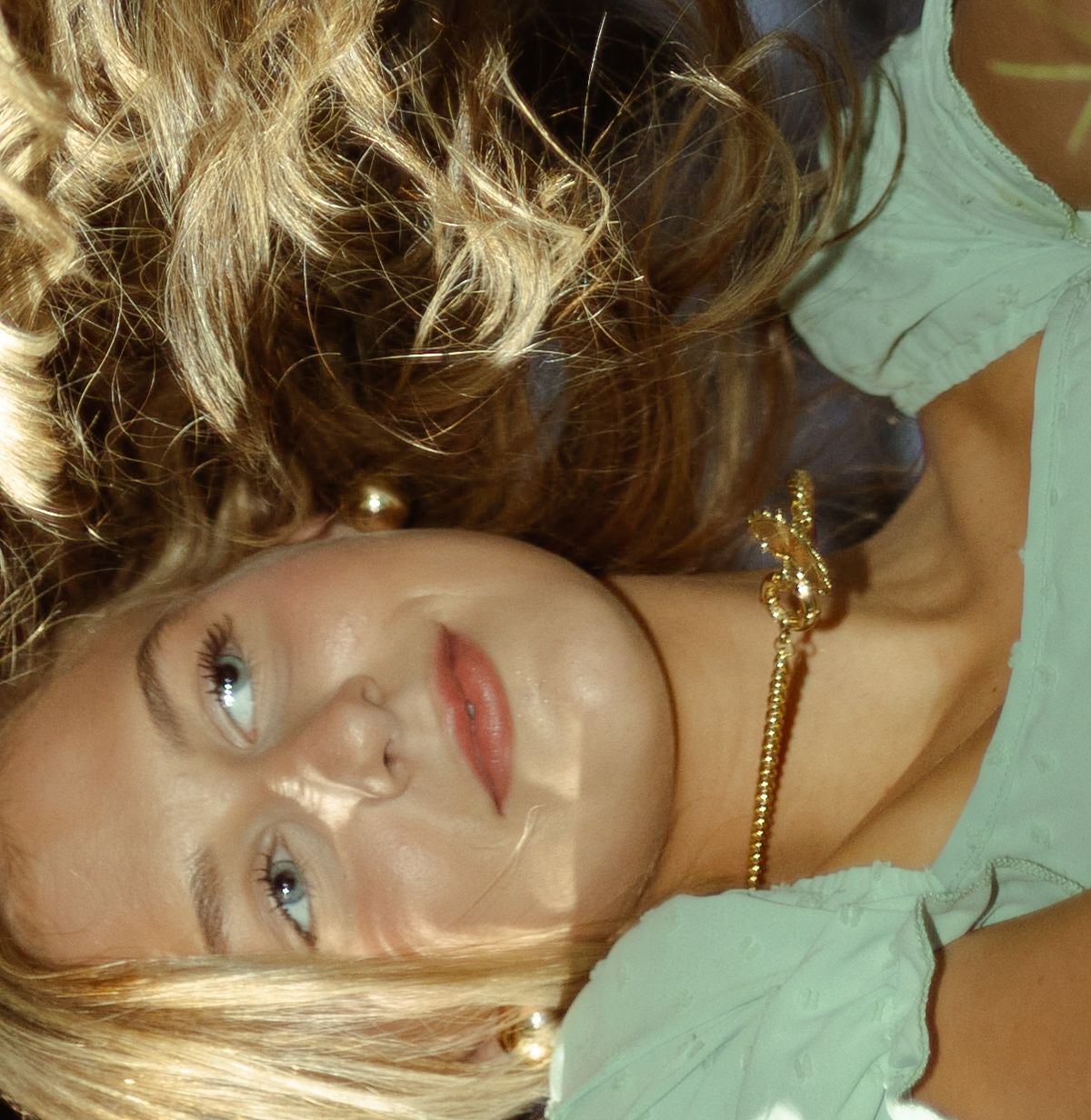 Close-up portrait of a blonde woman lying down wearing a gold bracelet and mint green top.