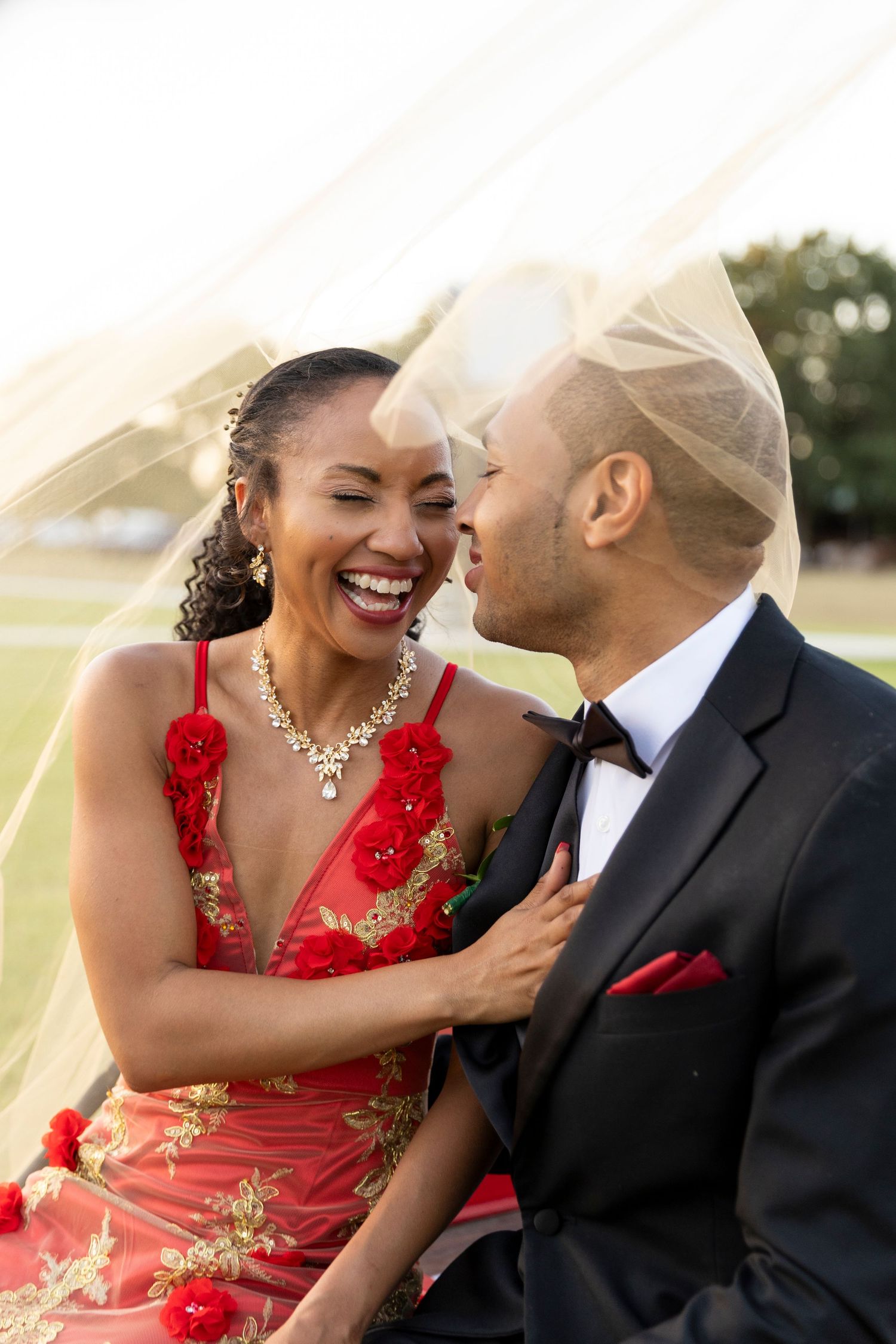 A laughing bride in a red floral gown and groom share a joyful moment under a wedding veil.
