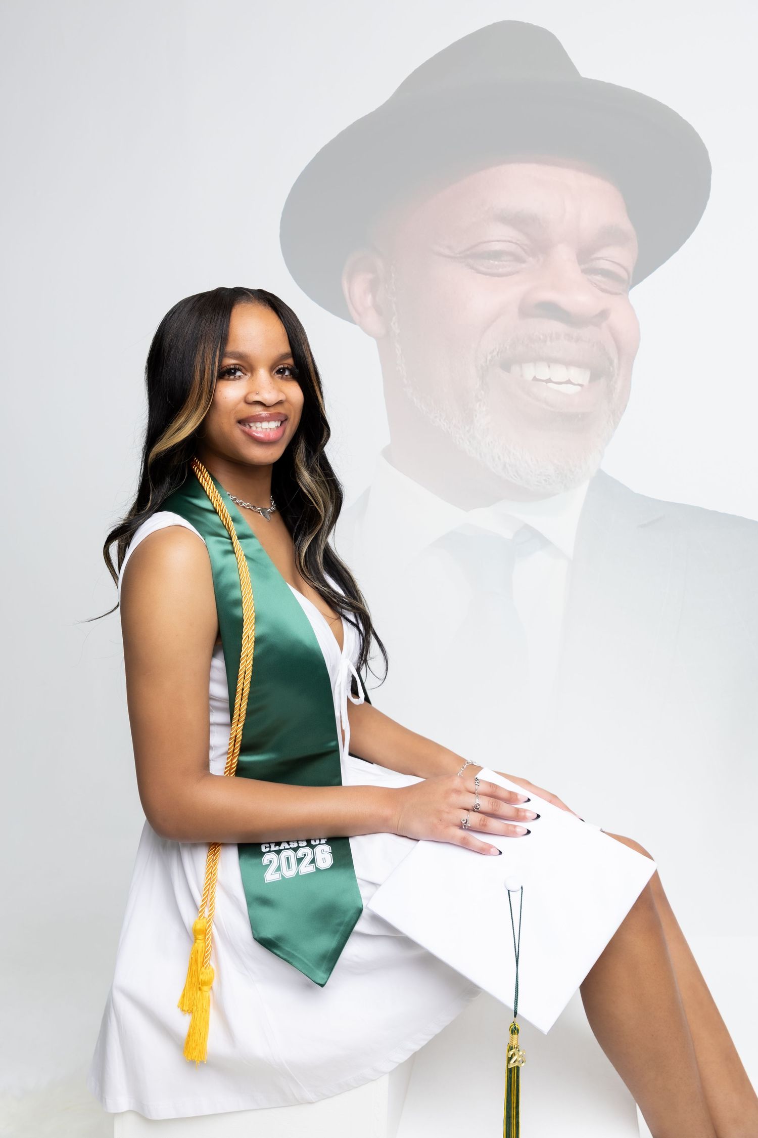 Smiling young Black woman in white dress with green graduation stole posing with diploma in studio.