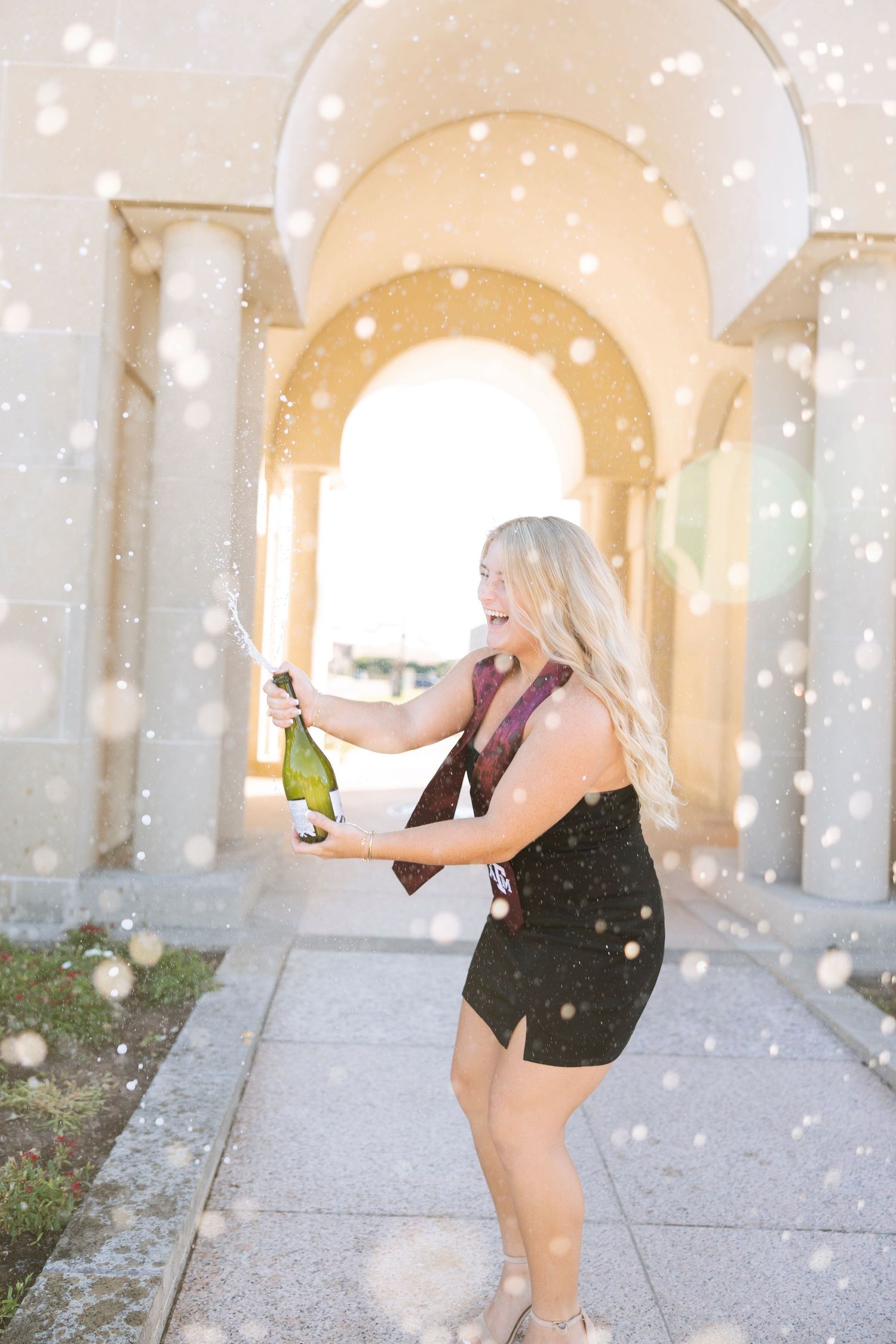 Blonde woman in black dress popping champagne bottle under arched columns with festive bokeh effect.