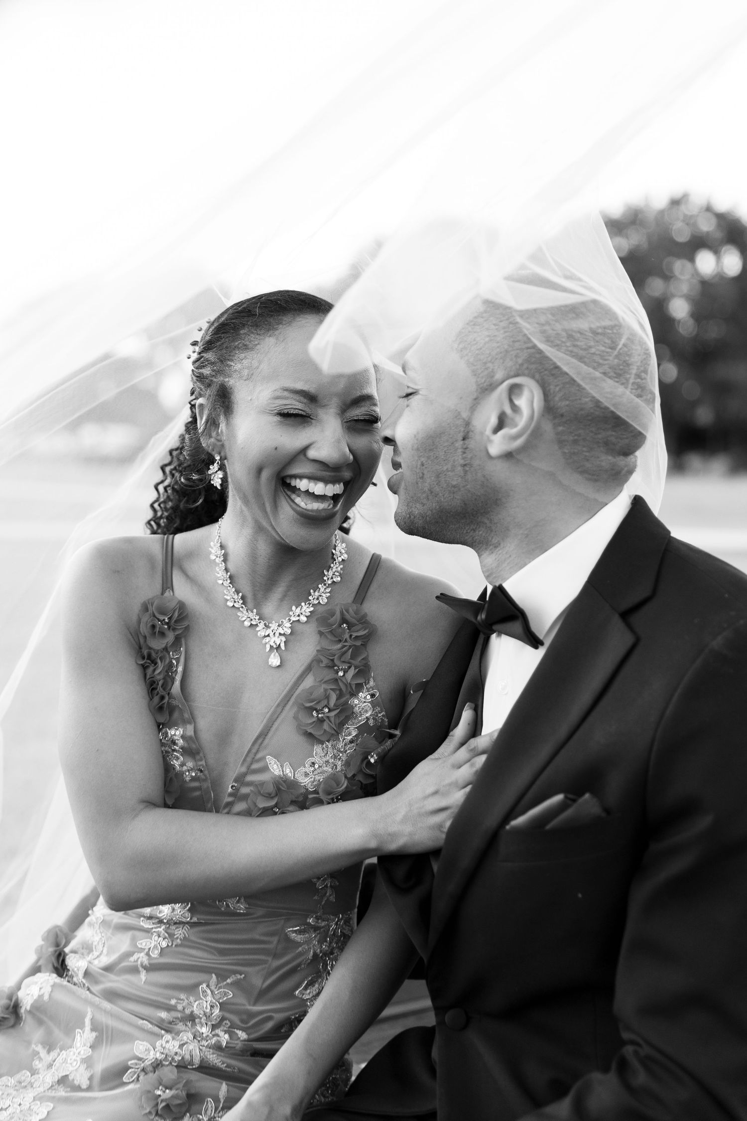 Black and white photo of a laughing bride and groom sharing a joyful moment under a flowing wedding veil.