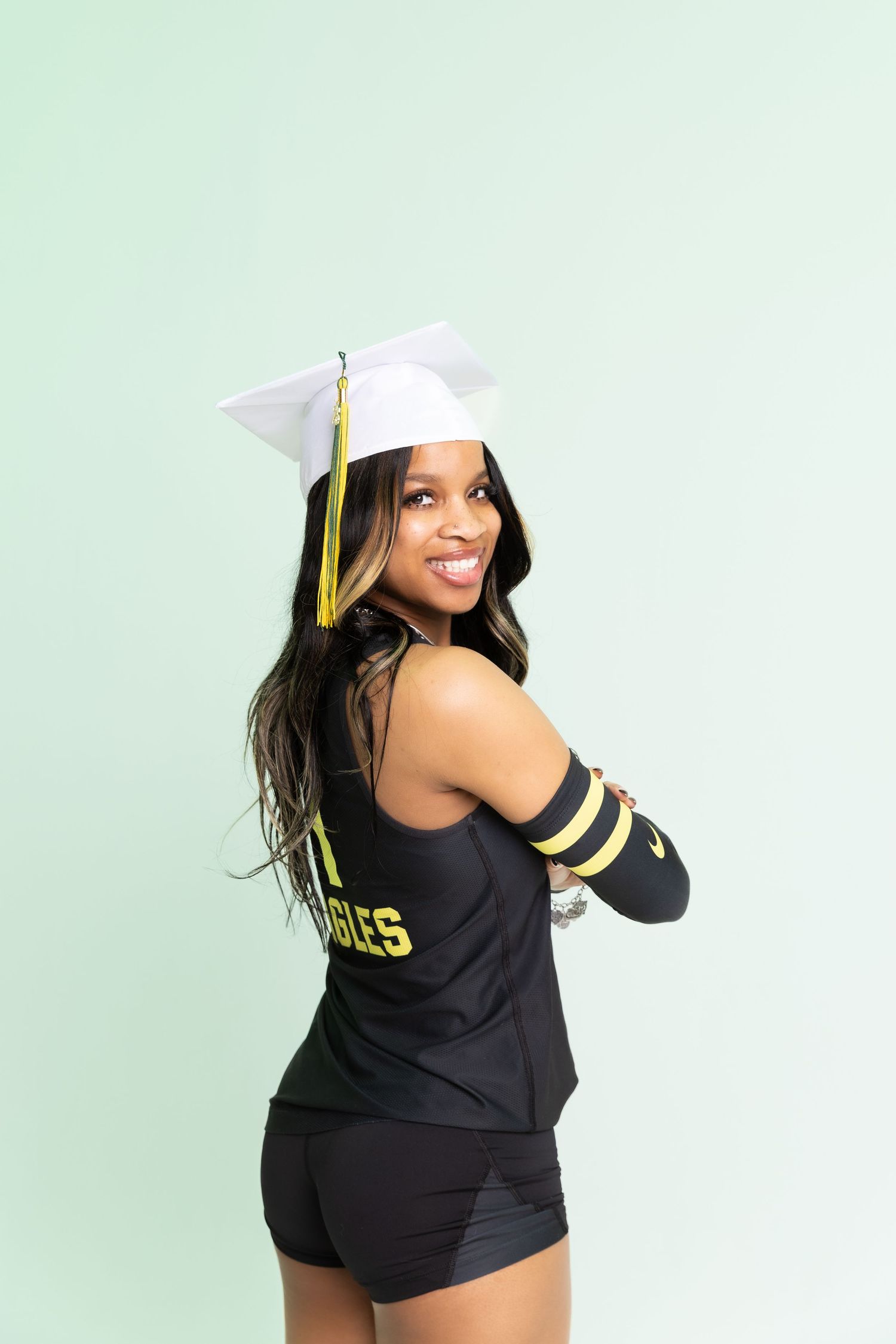 Smiling graduate in black jersey and white cap poses confidently against a mint green background.