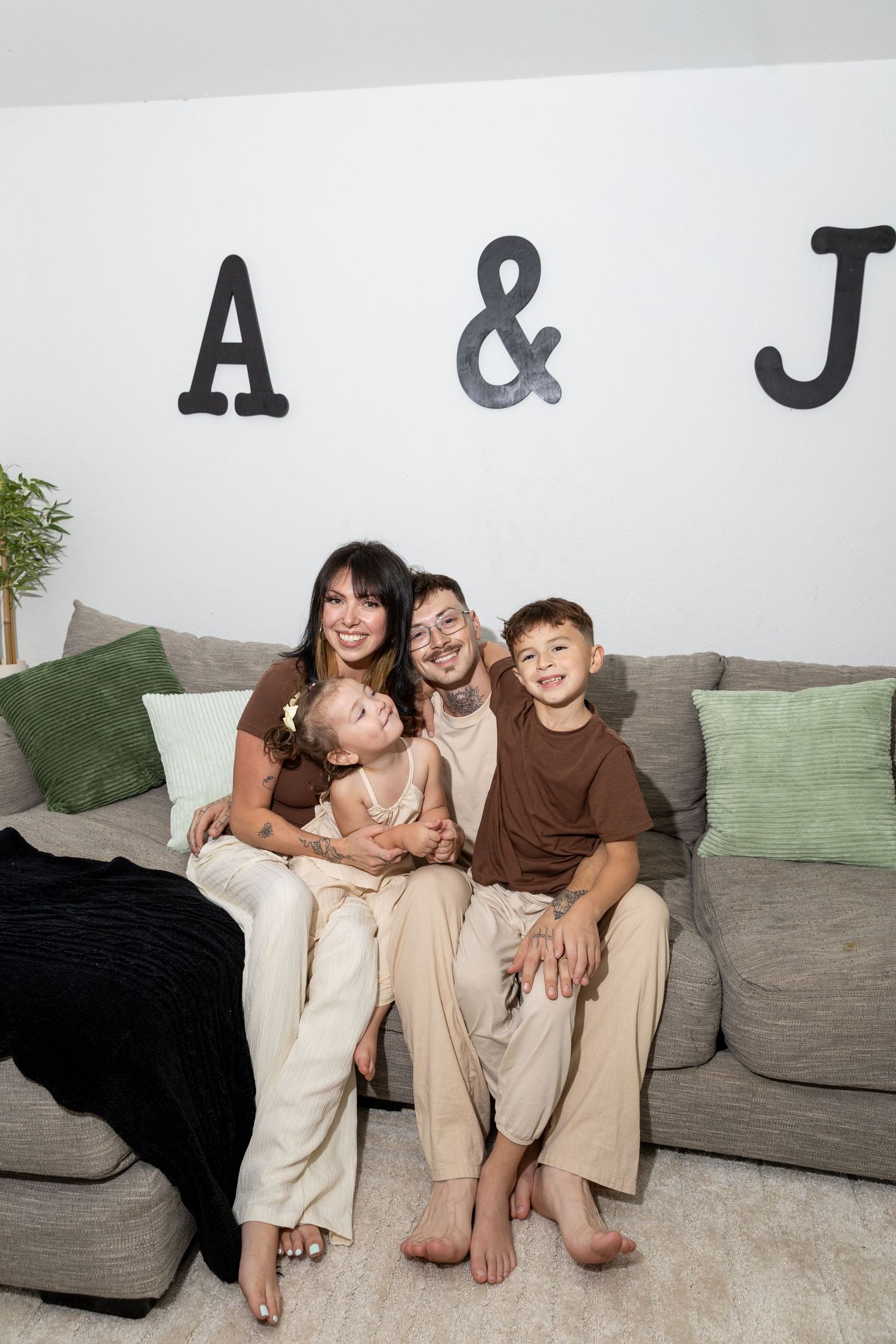 A happy family of four sits together on a gray couch beneath large letters A & J on a white wall.