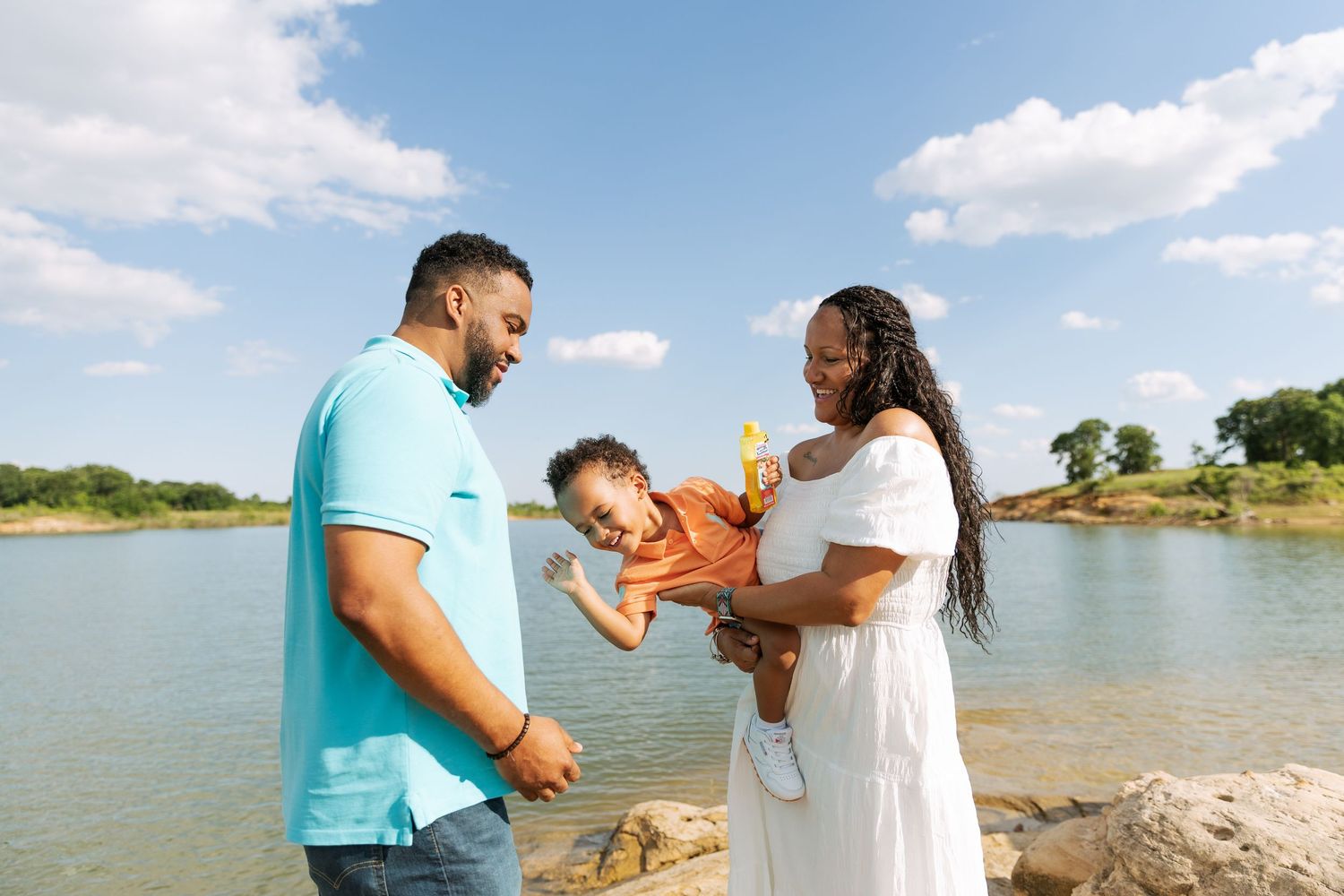Happy family of three enjoys sunny day by the lake with parents holding their young child.