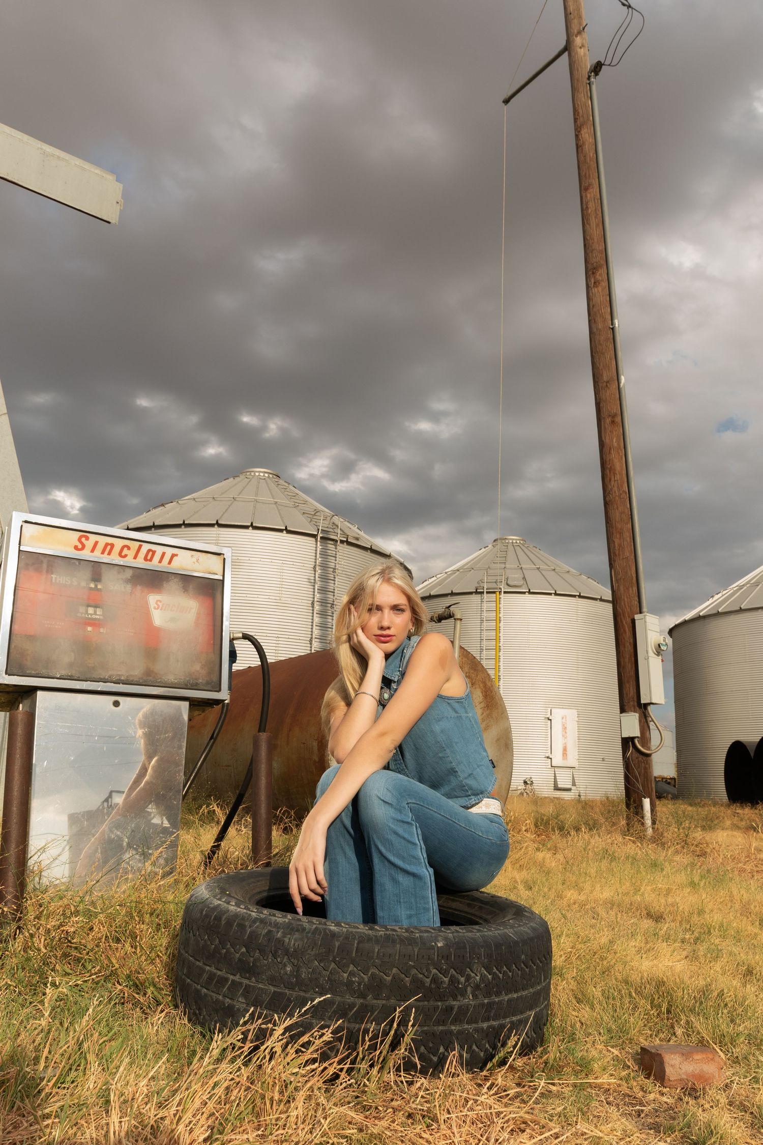 Blonde woman in denim overalls sitting on tires near grain silos and a Sinclair sign under stormy skies.