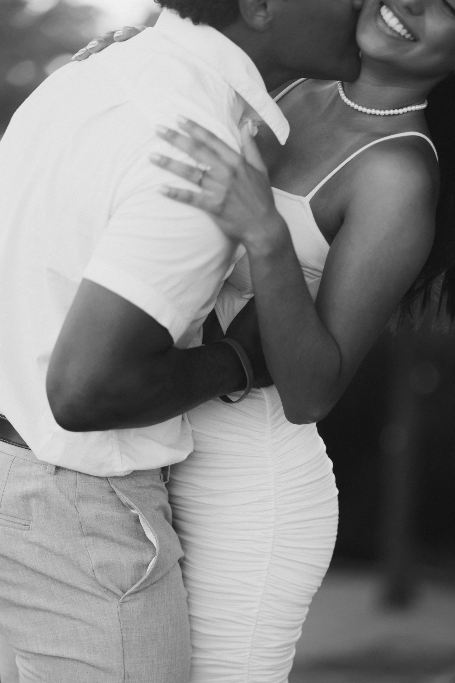 Black and white photo of a couple embracing, woman in white dress with pearl necklace smiling joyfully.