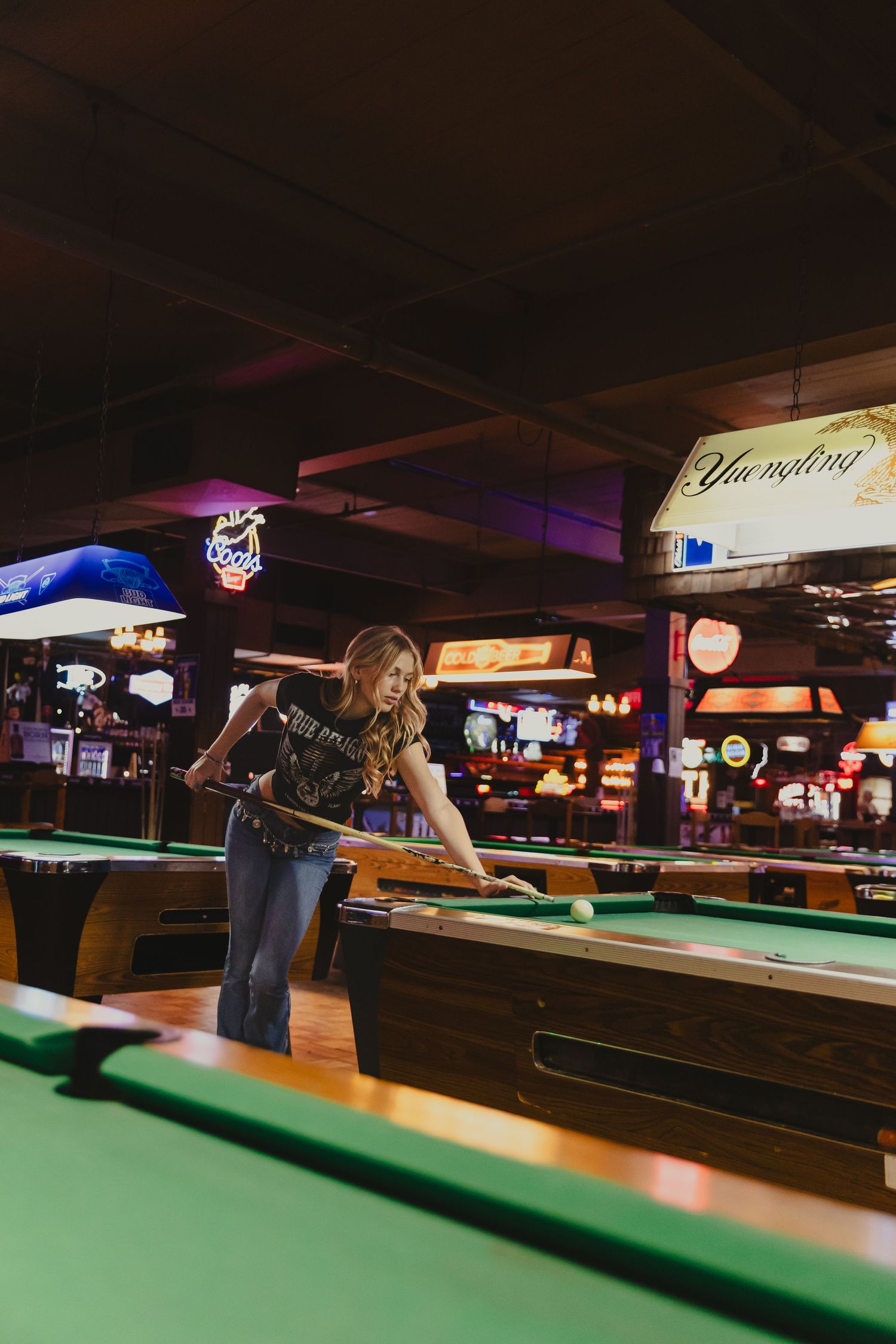 Woman playing pool at a bar with neon signs and Yuengling beer light in background.