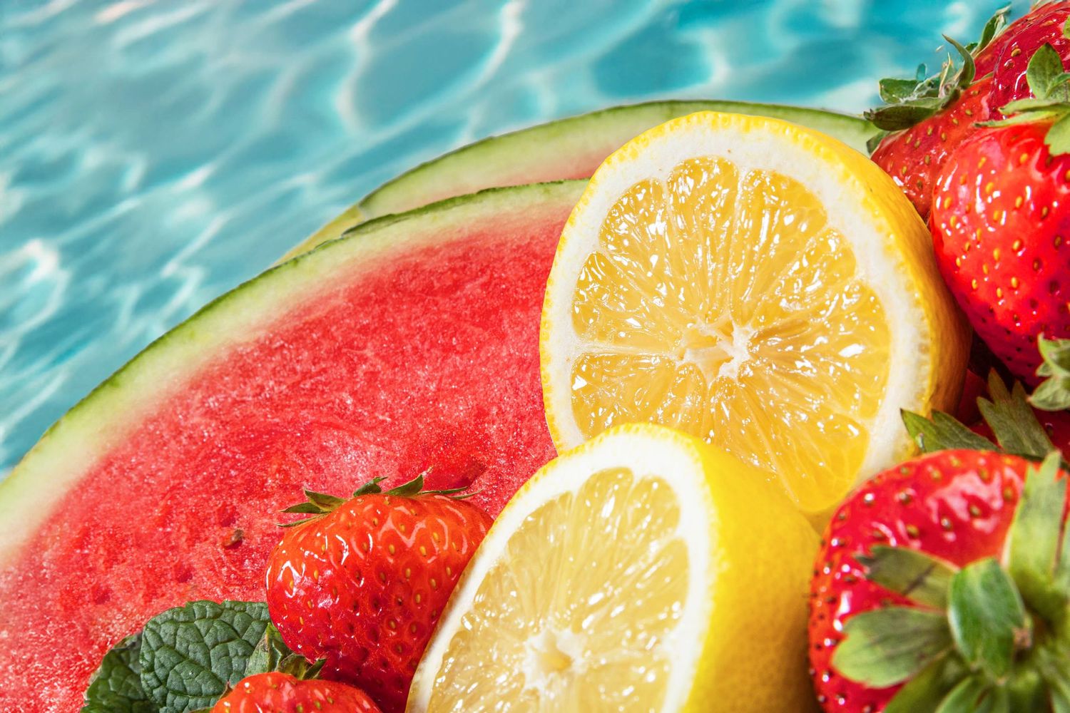 Fresh watermelon, lemon slices, and strawberries floating by a bright blue pool on a sunny summer day.