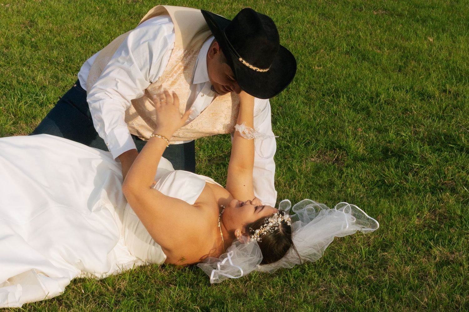 Bride in white wedding gown lying on green grass while groom in cowboy hat leans down to kiss her.