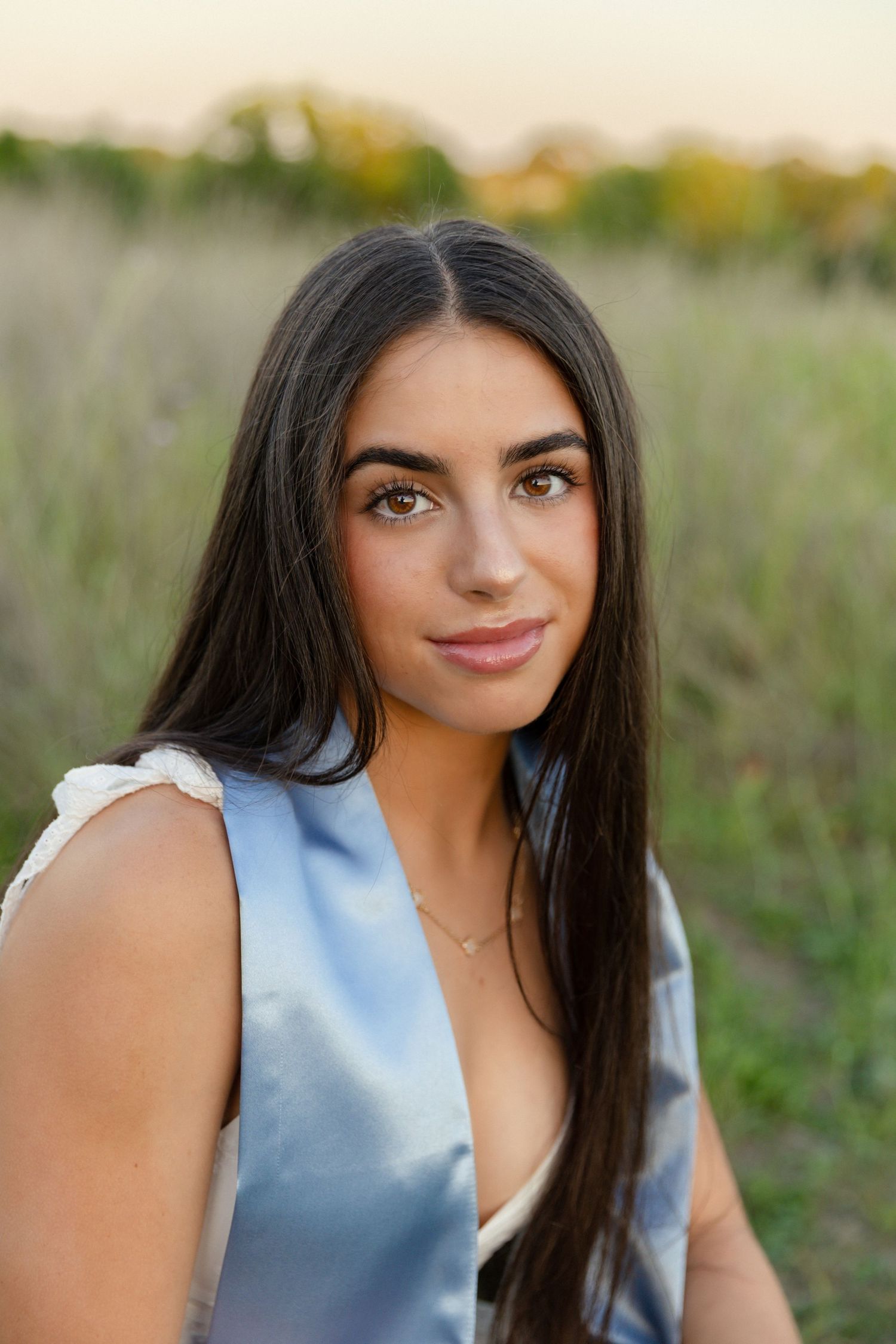 Young woman with long dark hair in a blue sleeveless dress poses in a golden field at sunset.