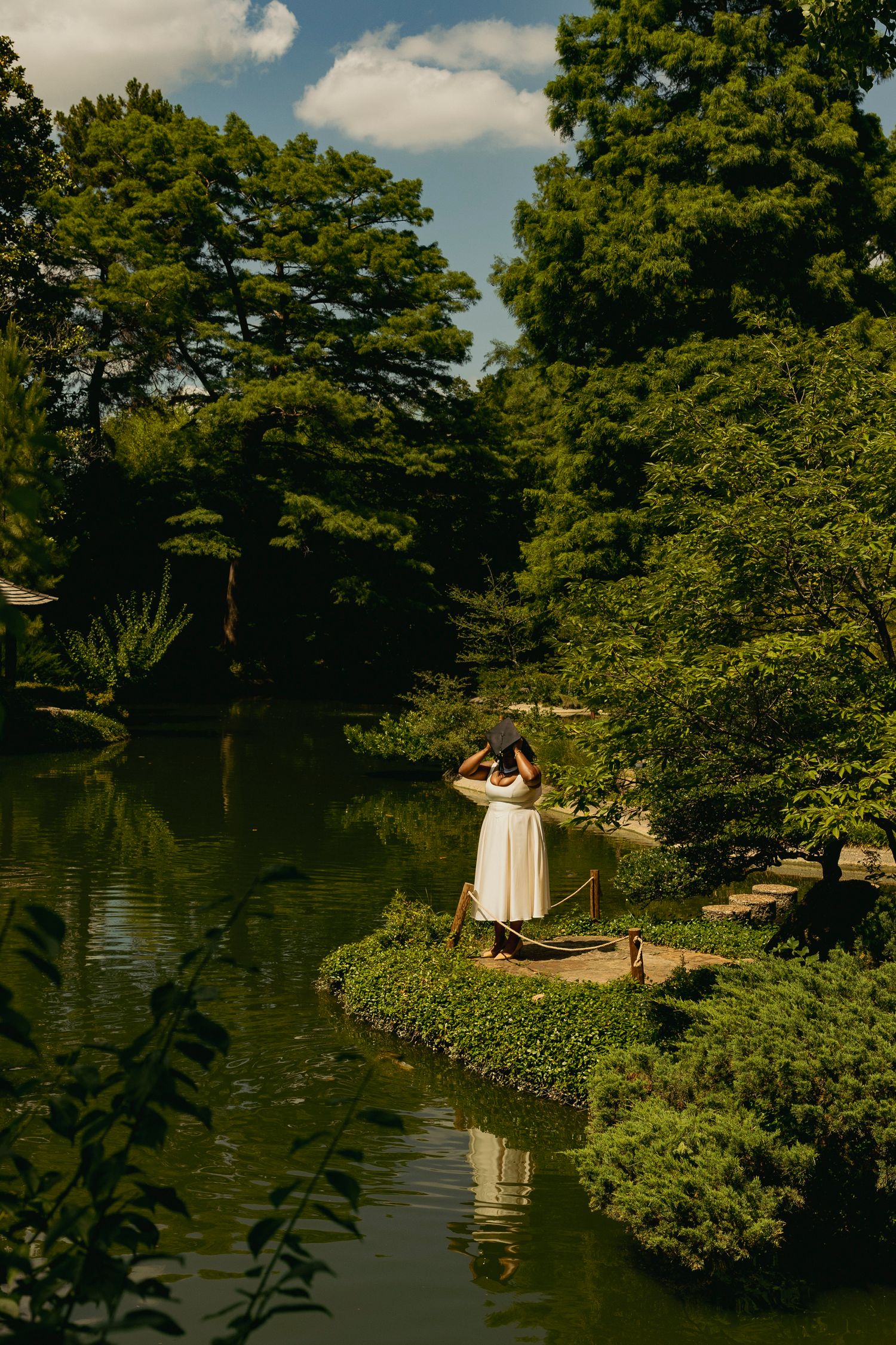 Woman in white dress stands on garden island surrounded by lush green trees beside a serene pond.