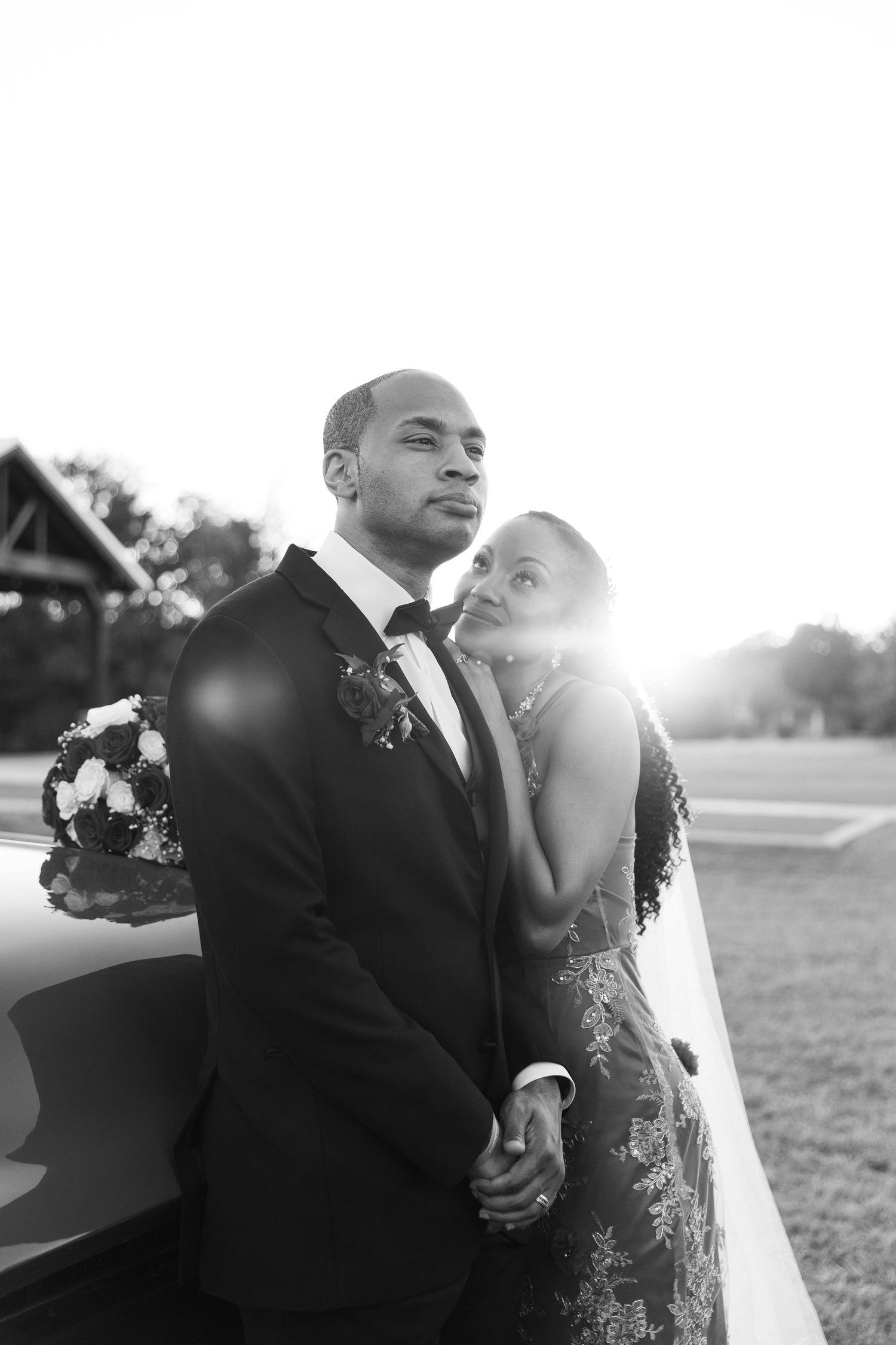 A black and white photo of a bride and groom holding hands and leaning against a vintage car at sunset.