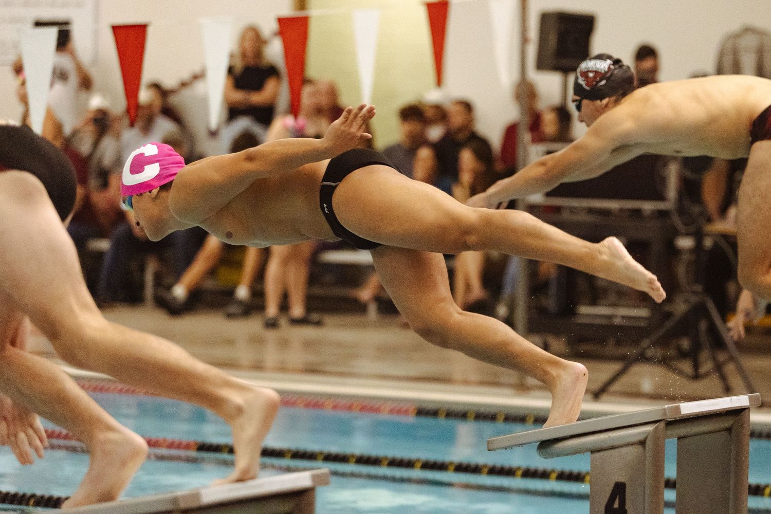 Competitive swimmers in pink caps diving off starting blocks at indoor swim meet.