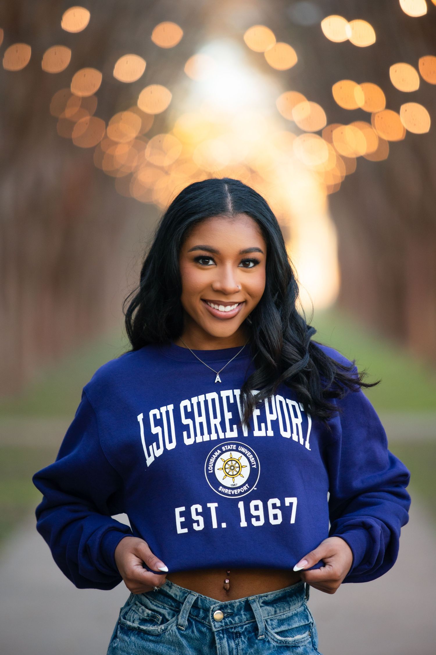 Young woman smiling in an LSU Shreveport EST. 1967 blue cropped sweatshirt with bokeh lights background.