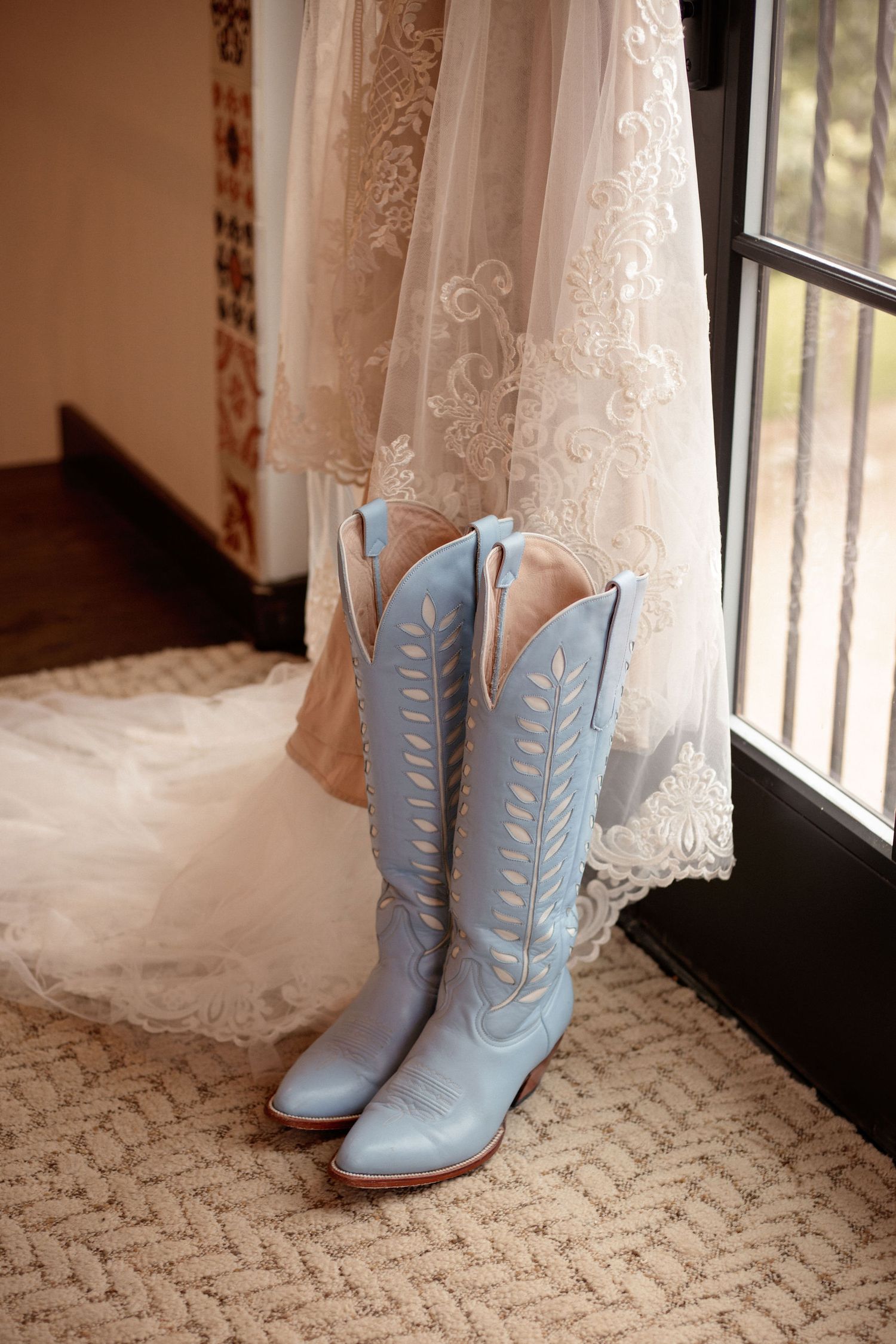 Western-inspired wedding details featuring a lace wedding dress and sky blue cowgirl boots in natural window light.