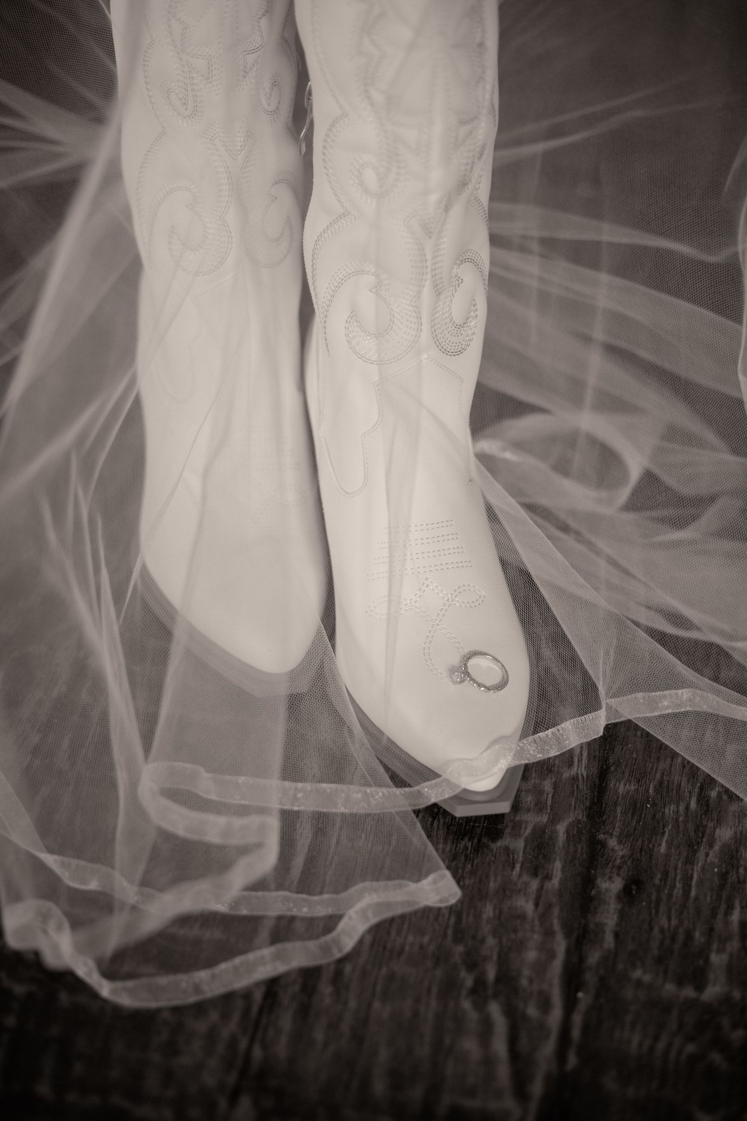 Black and white documentary wedding photo of a wedding band resting on a cowboy boot beneath a flowing bridal veil.