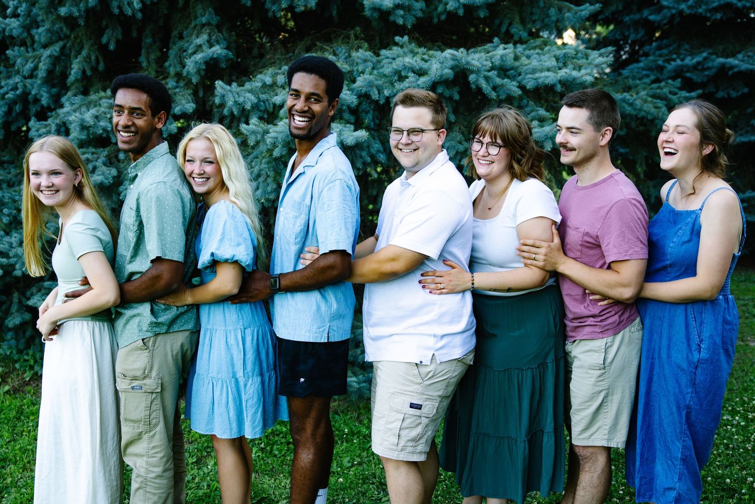 Group of eight young adults smiling and laughing together outdoors in front of green trees on a sunny day.