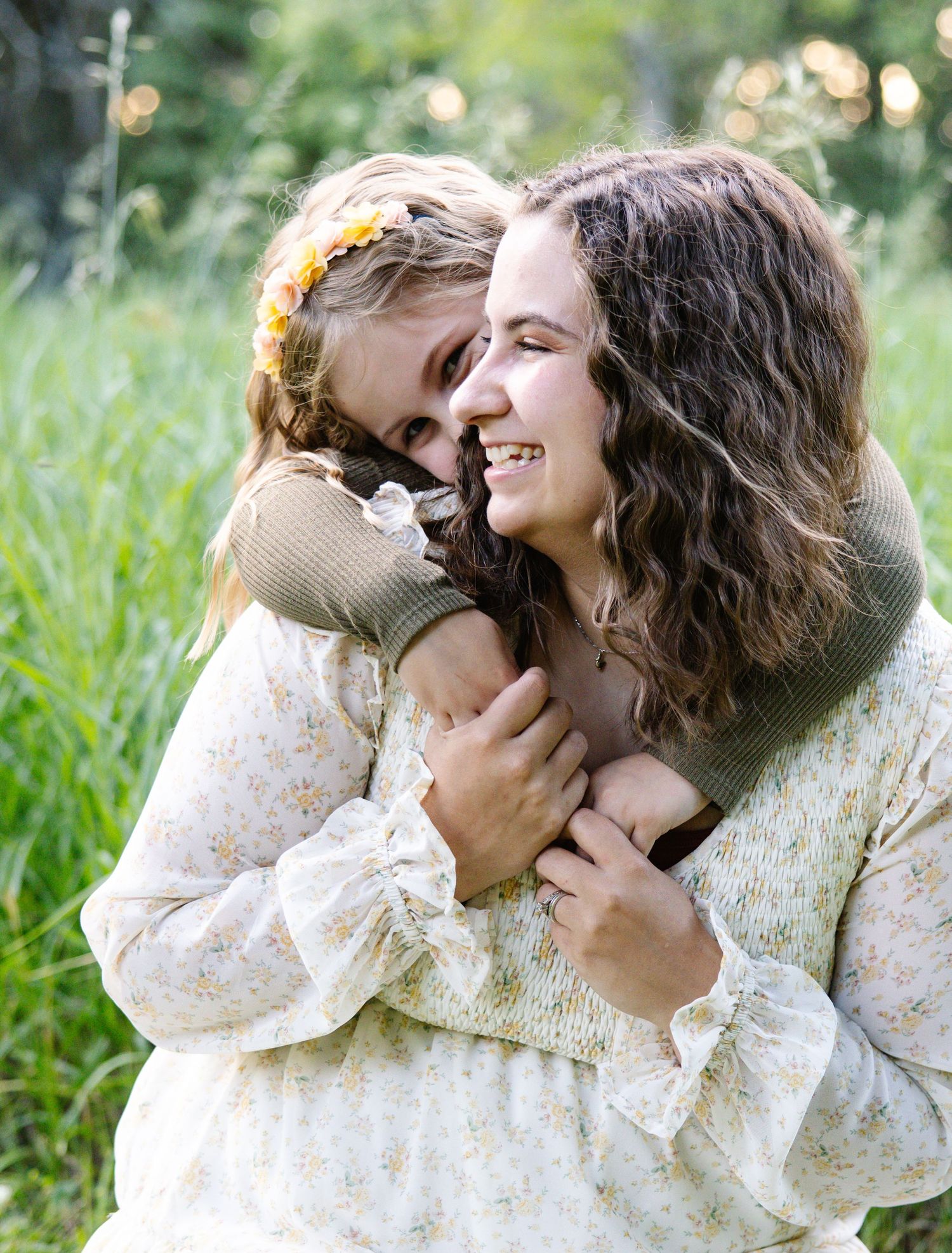 A smiling mother embraces her laughing daughter outdoors in a green meadow, both wearing floral outfits.