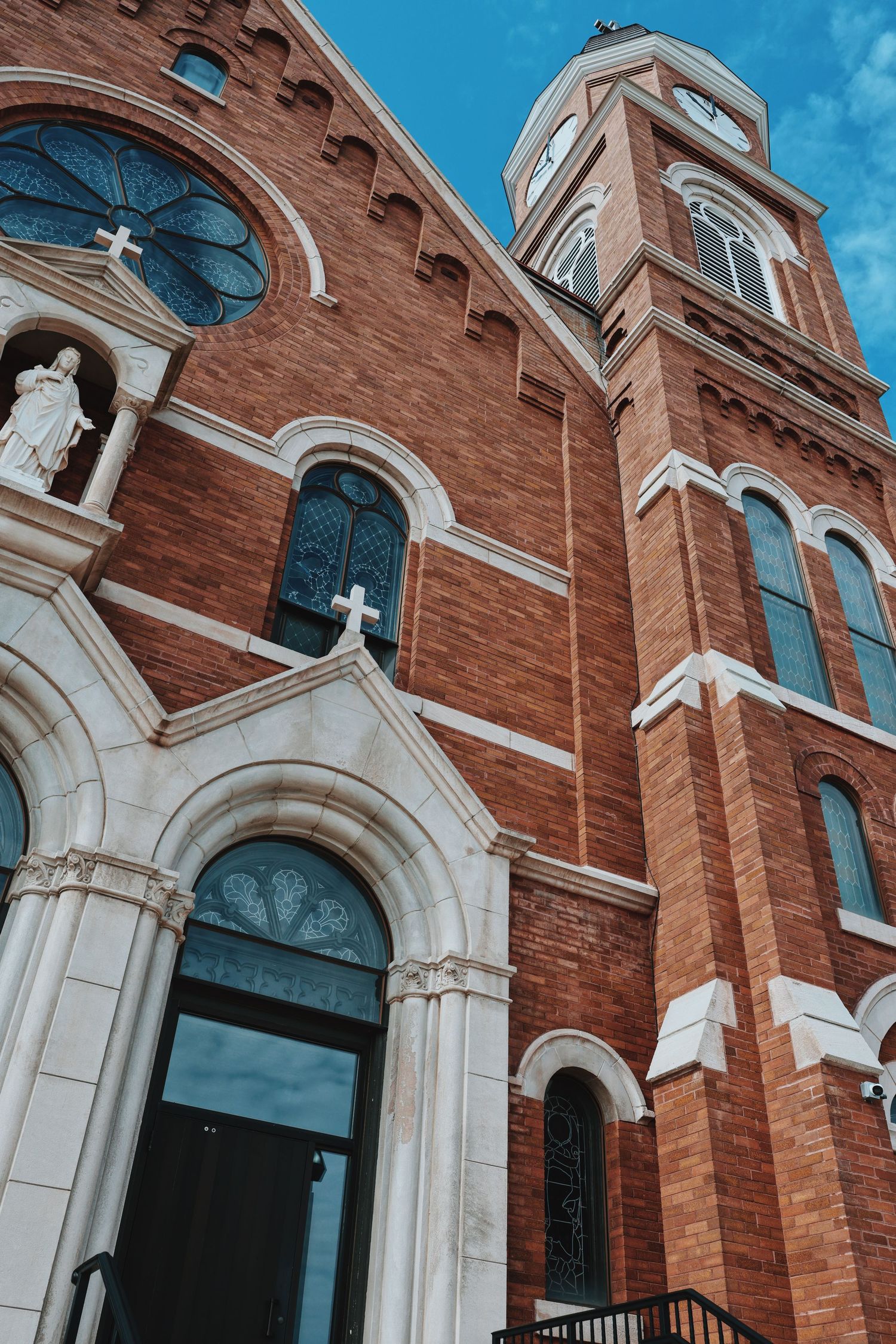 Red brick Catholic church facade with arched windows, white stone trim, crosses, and a tall bell tower against blue sky.