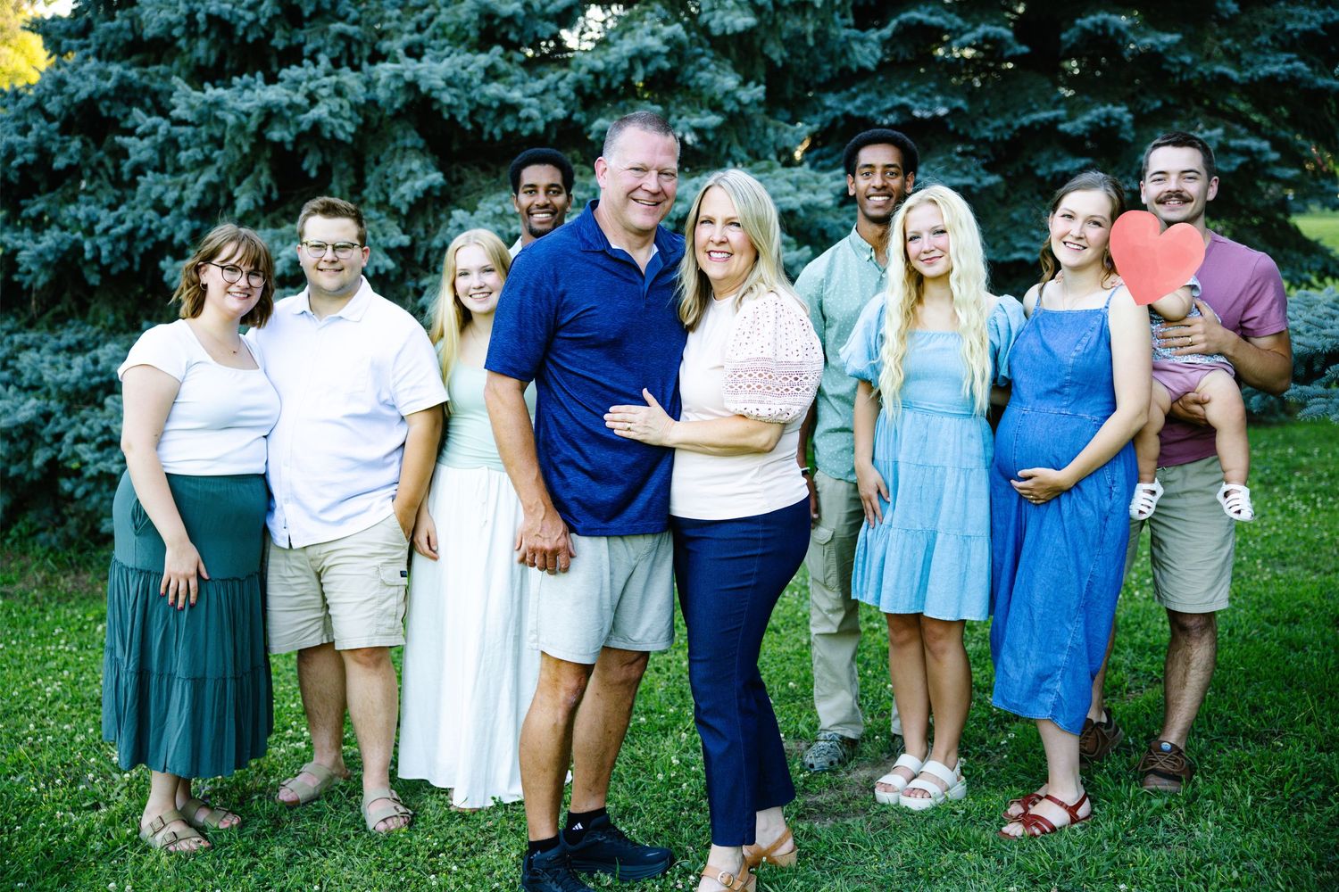 Large family group portrait smiling outdoors on green lawn with blue spruce trees in background on sunny day.