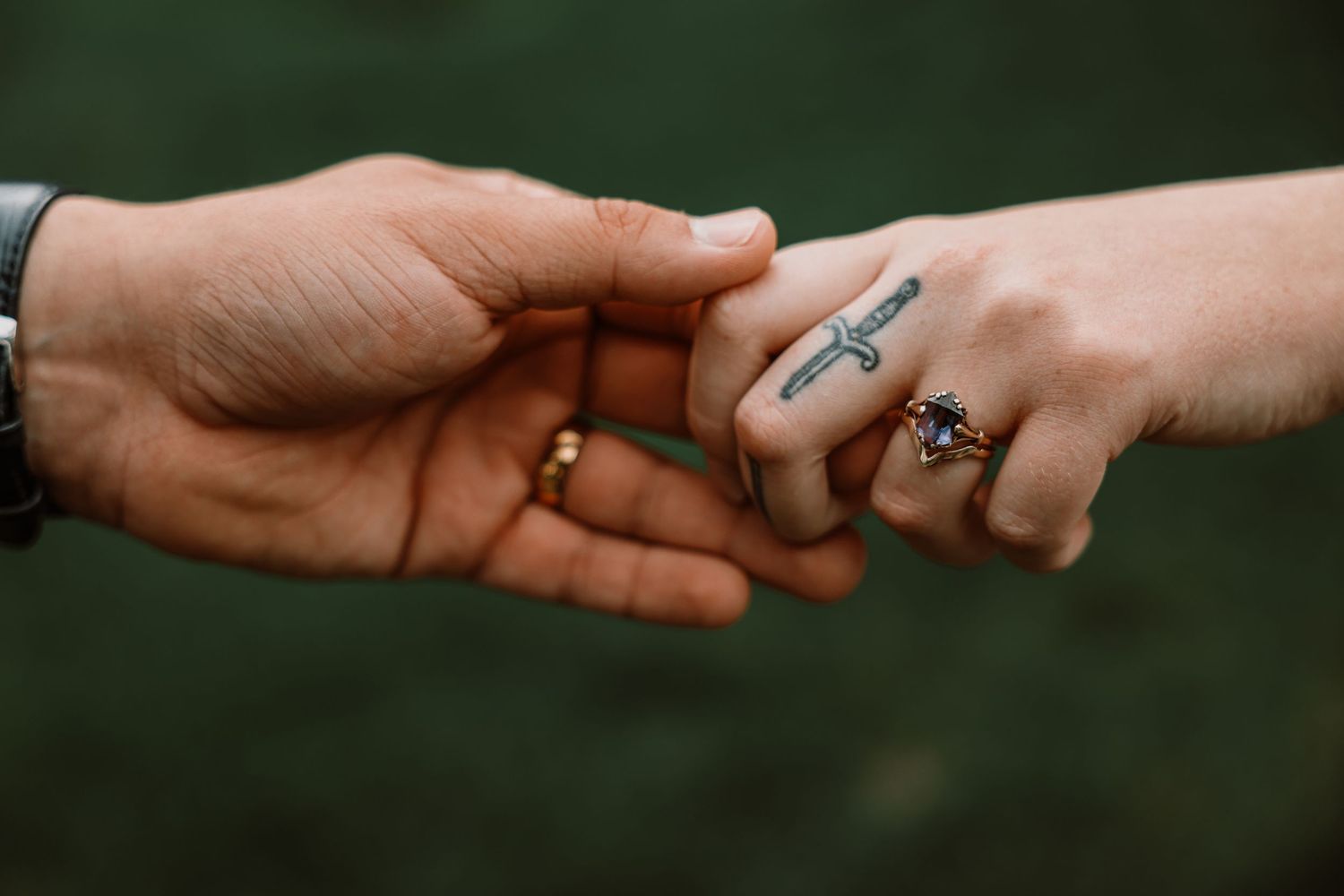 Two hands gently linking pinkies, one with a cross tattoo and gemstone ring, symbolizing love and connection.