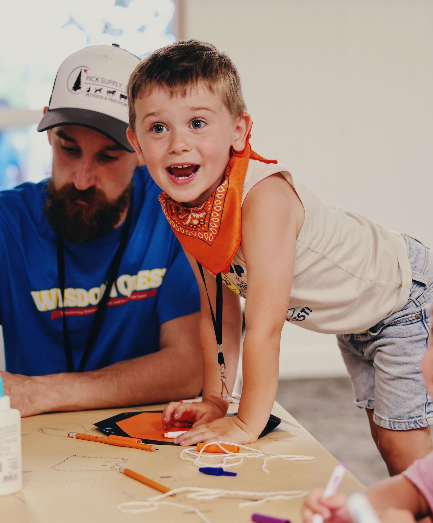 A young boy in an orange bandana smiles at a craft table with a bearded man in a blue shirt at summer camp.