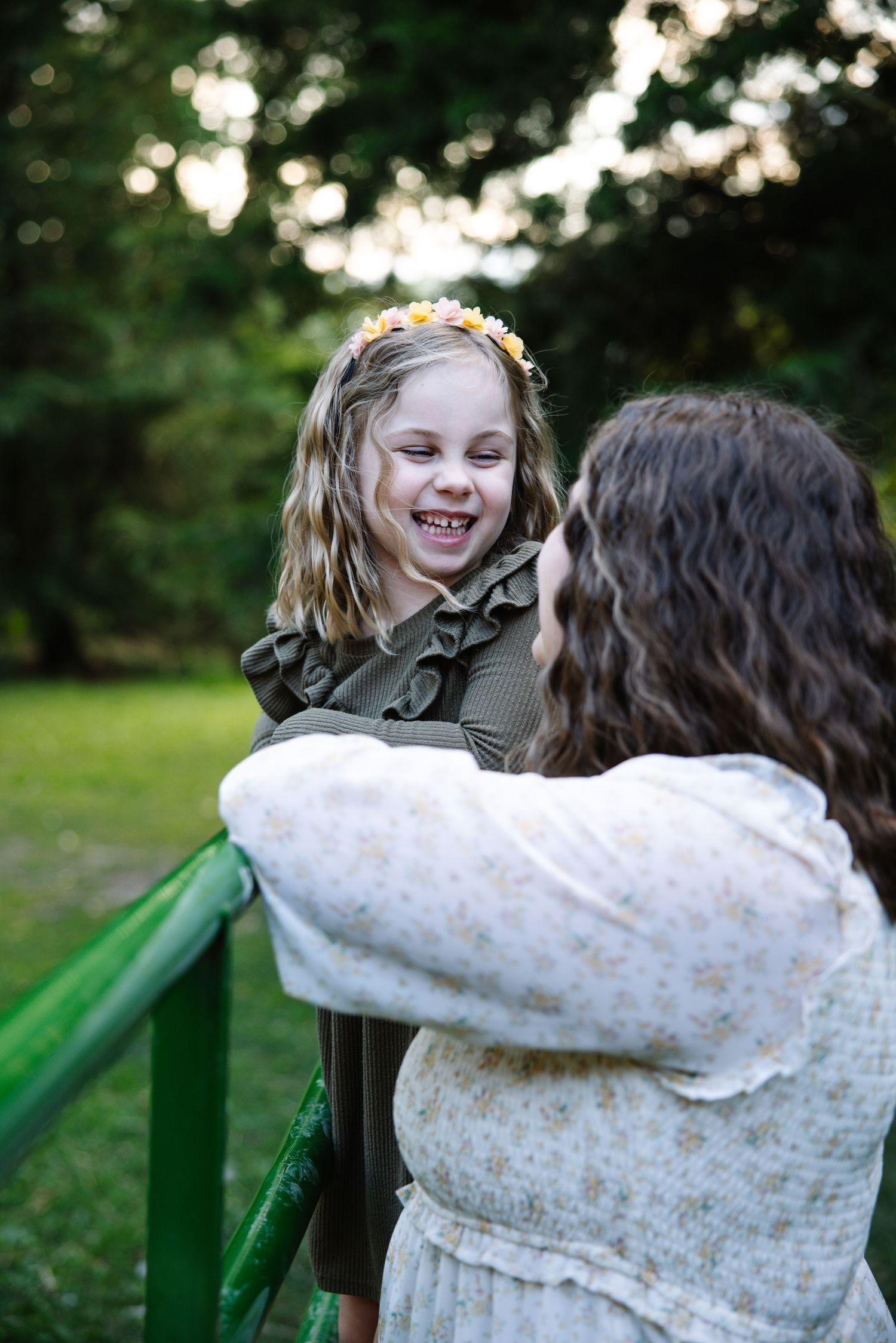 A joyful young girl wearing a floral crown laughs while a curly-haired woman in a floral dress holds her near a green railing.