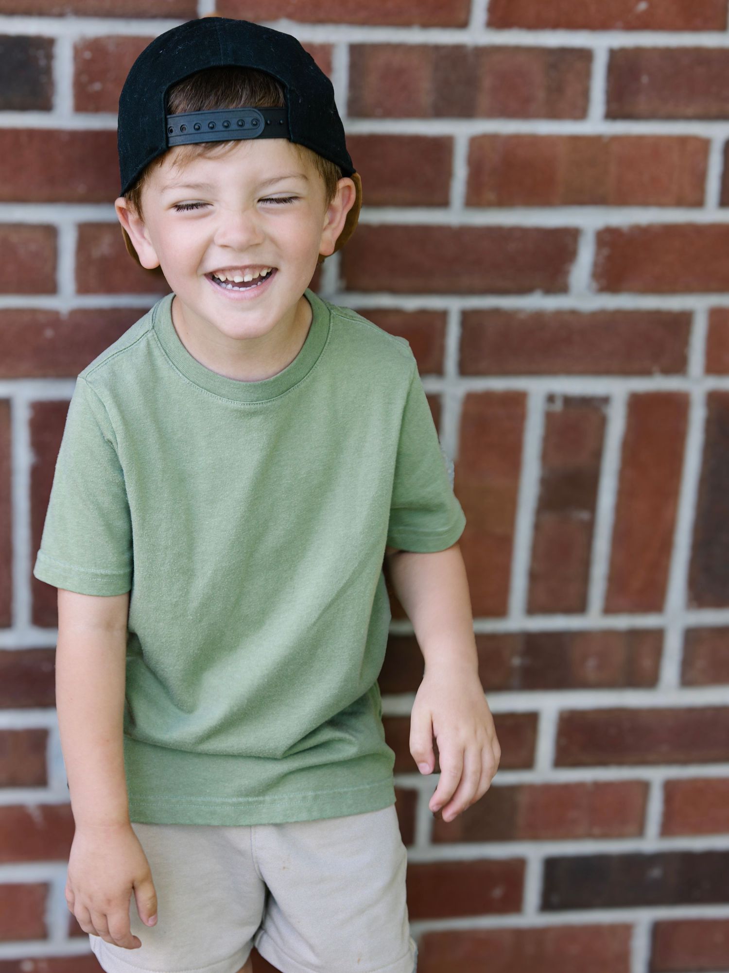A smiling boy in a green t-shirt and backwards black cap poses happily against a red brick wall.