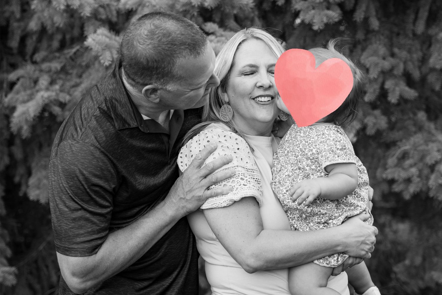Black and white photo of a couple sharing a tender moment, woman holding a child with a red heart emoji covering the child's face.