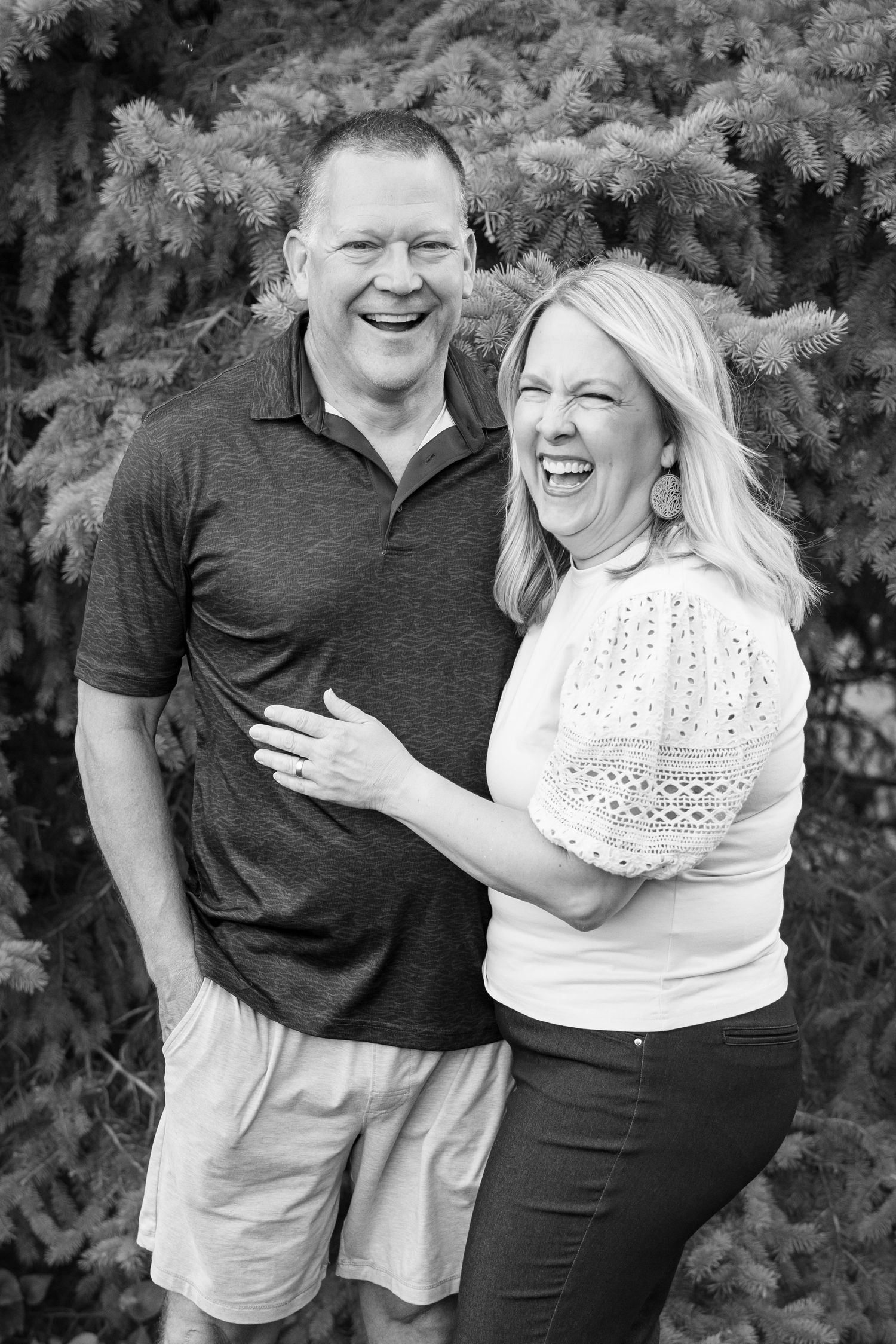 Black and white photo of a laughing couple posing outdoors in front of evergreen trees.