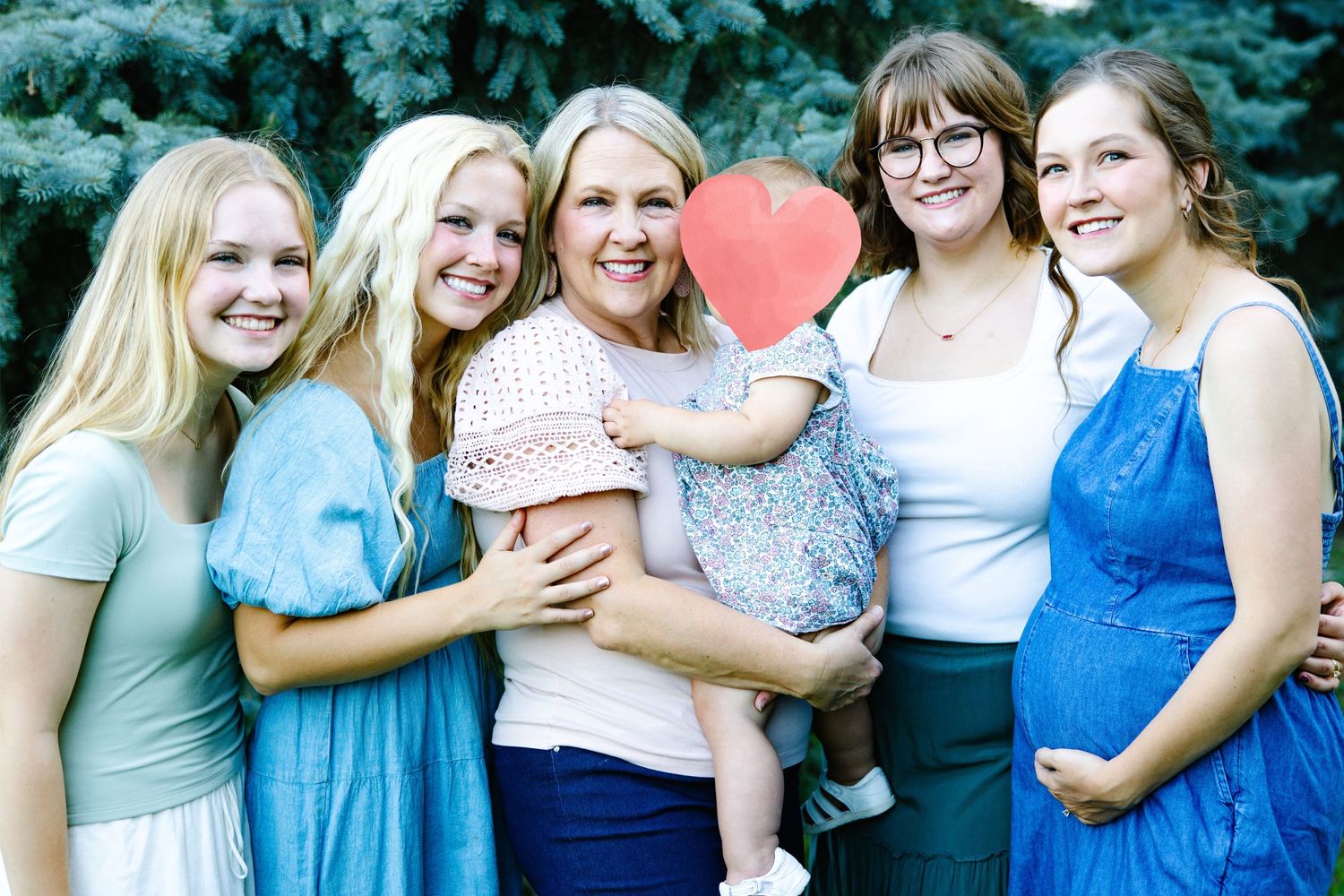 Five women smiling outdoors near evergreen trees, with one holding a baby covering its face with a heart.