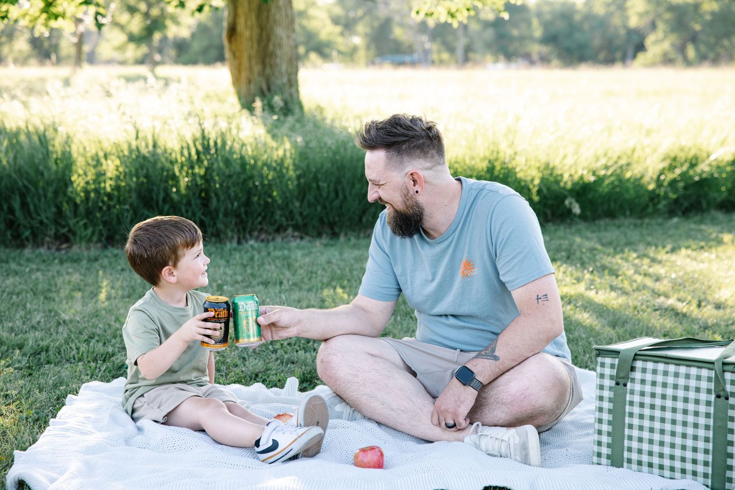 Father and young son enjoy a picnic in a sunny park, sitting on a blanket and sharing drinks together.