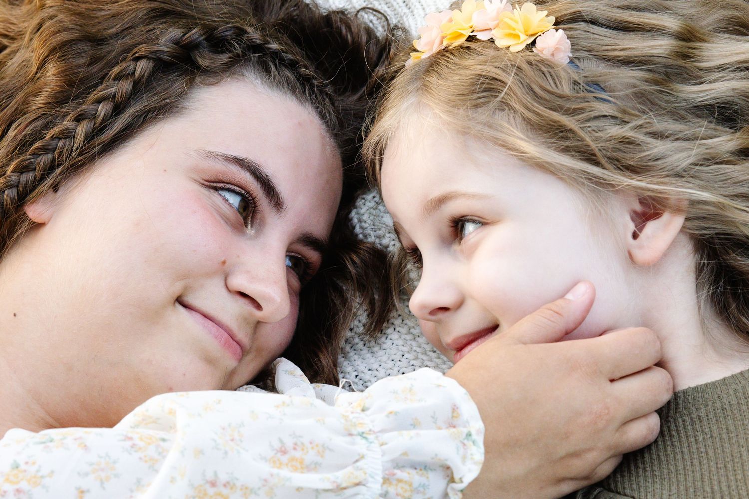 Two girls lying down face to face, older girl smiling warmly at younger girl wearing flower hair accessory.