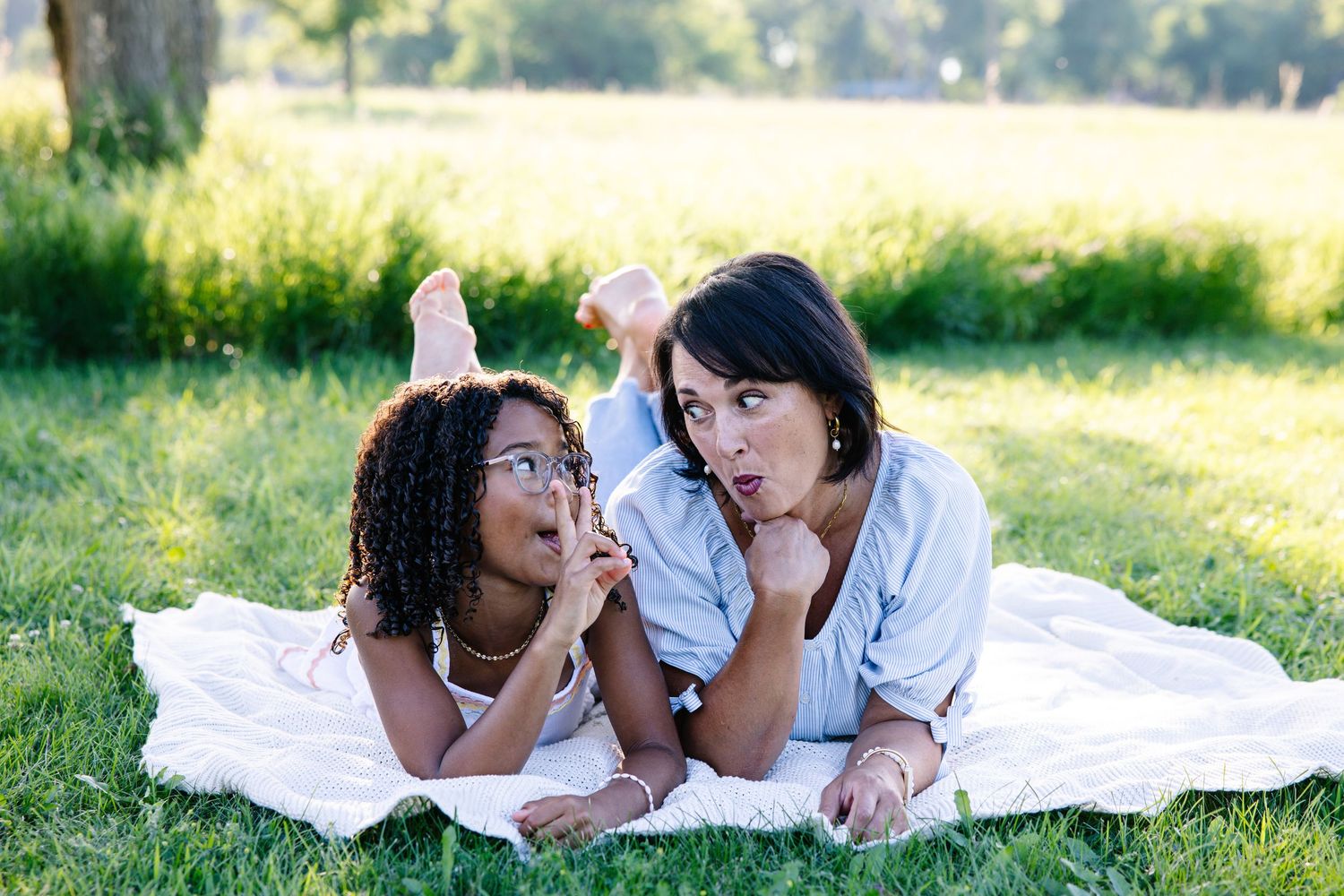 A woman and young girl lying on a blanket in a park, sharing a joyful moment together outdoors.