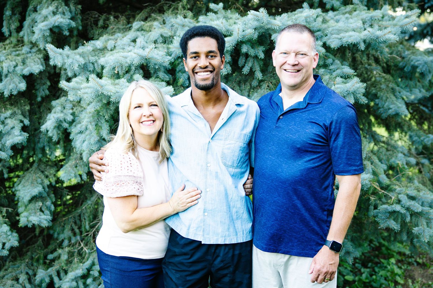 Smiling family of three posing outdoors in front of a blue spruce tree on a sunny day.