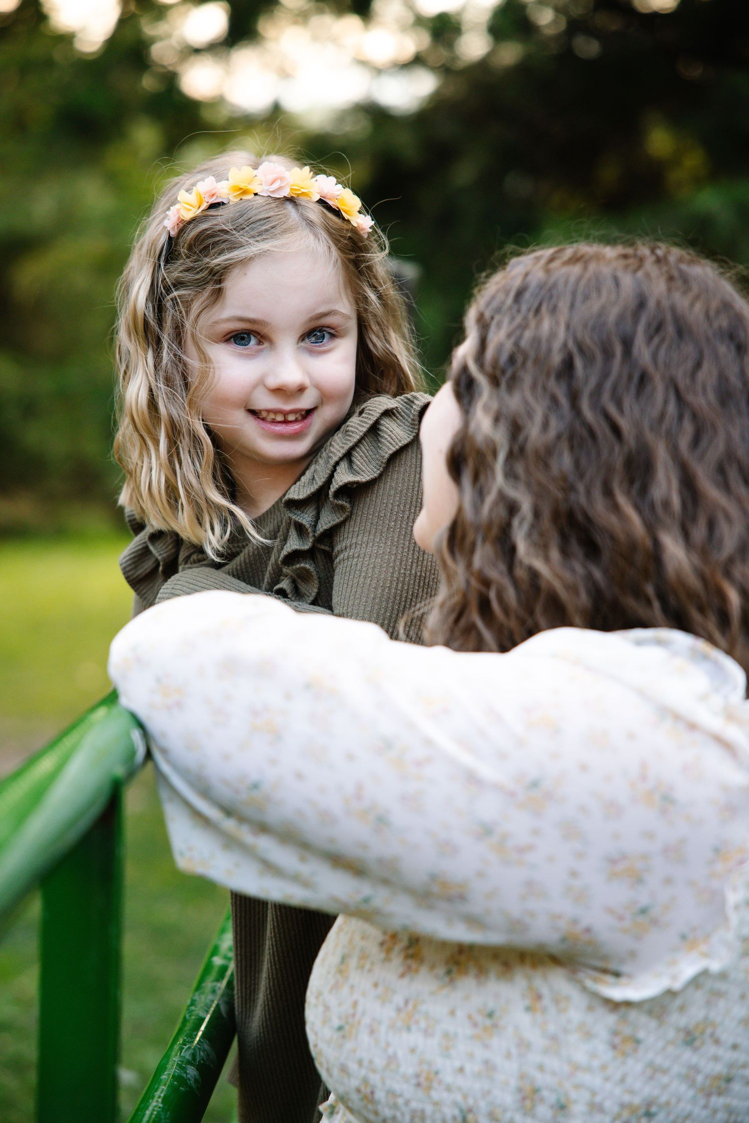 A young girl with a flower crown smiles at her curly-haired mother outdoors.