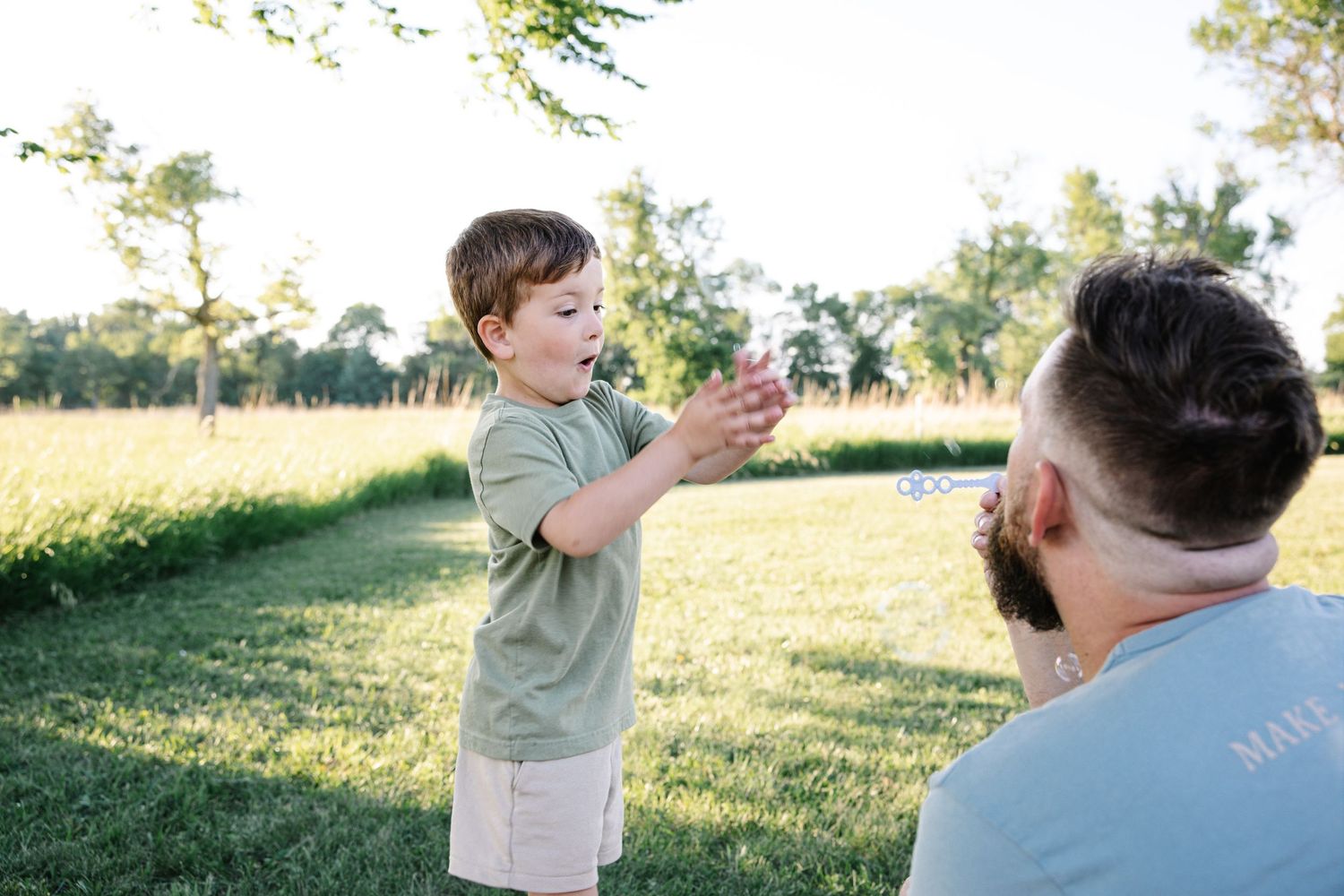 A happy toddler plays with bubbles with his bearded father in a sunny green park.