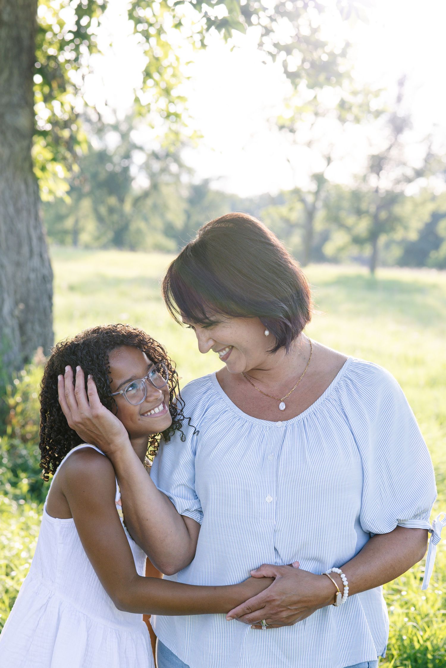 A woman and young girl in white dresses share a joyful embrace outdoors in a sunny green park.