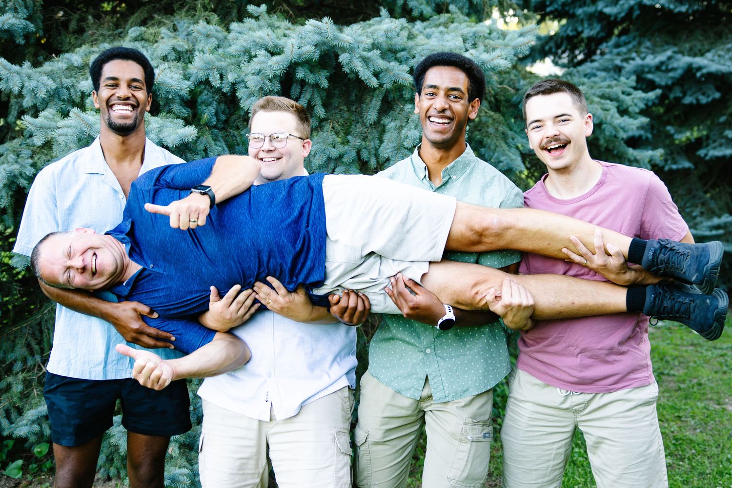 Five young men laughing outdoors, four friends carrying one man horizontally in front of green trees.