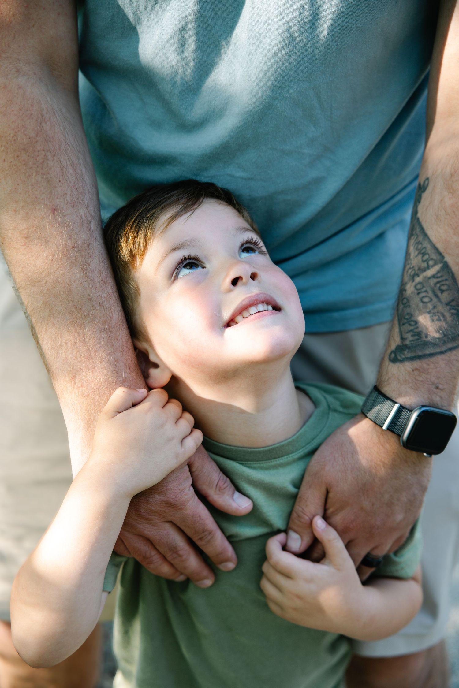A young boy smiles up at his father while being held in a warm embrace outdoors.