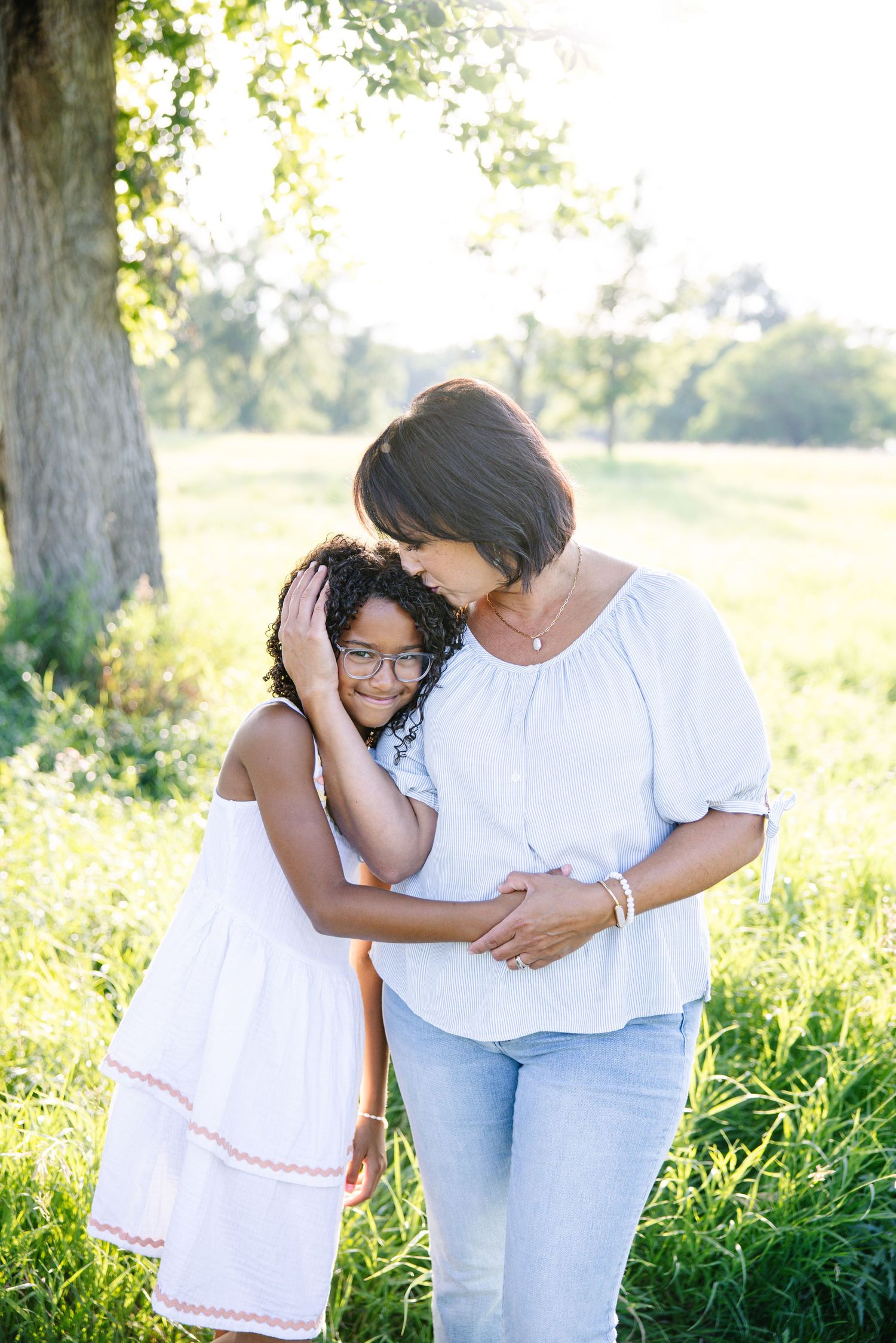 A woman in white top and jeans tenderly embraces a young girl in a white dress outdoors in a sunny green park.