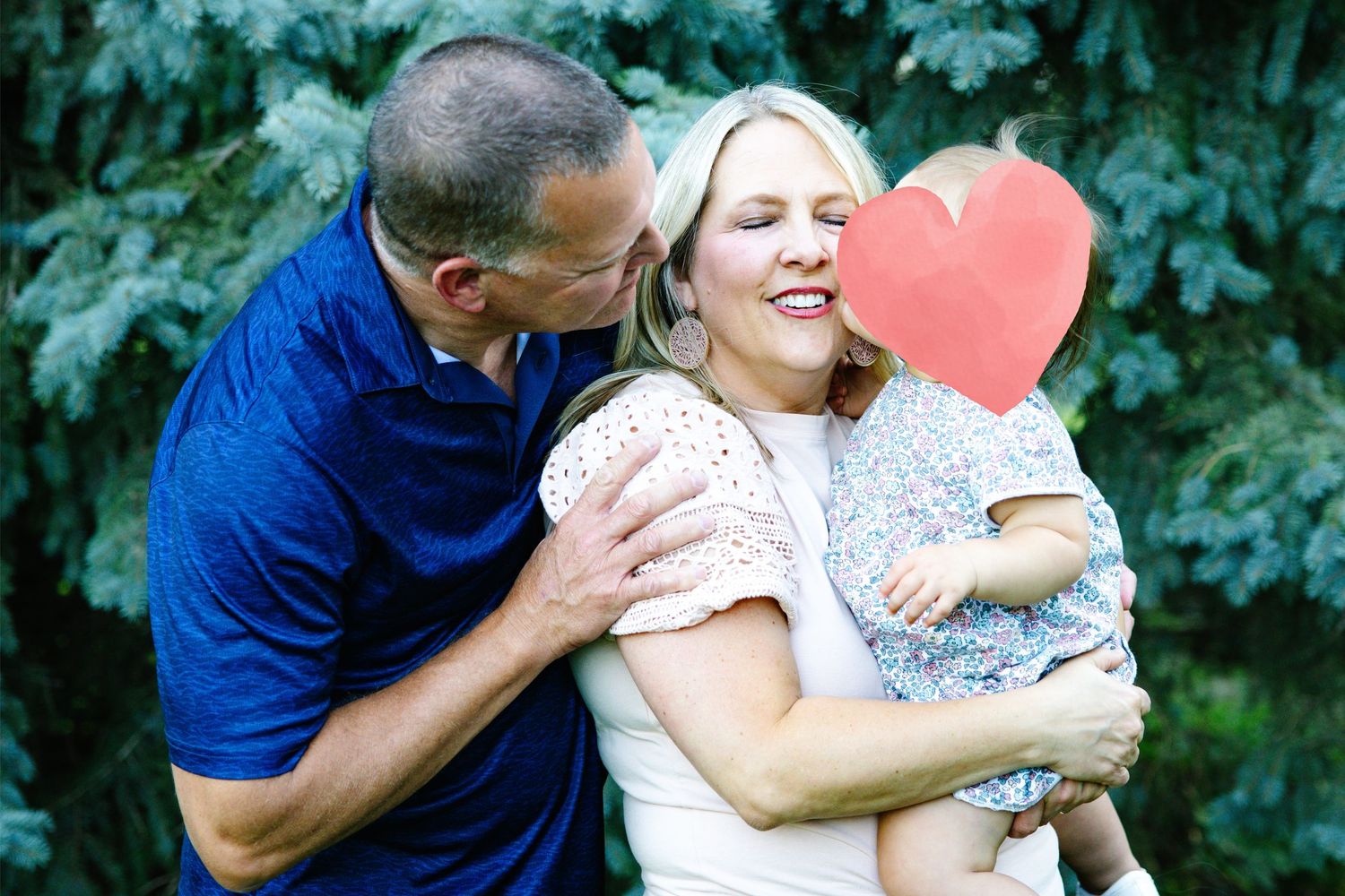 A happy family with a man kissing a smiling woman holding a baby with a red heart cutout covering the child's face.