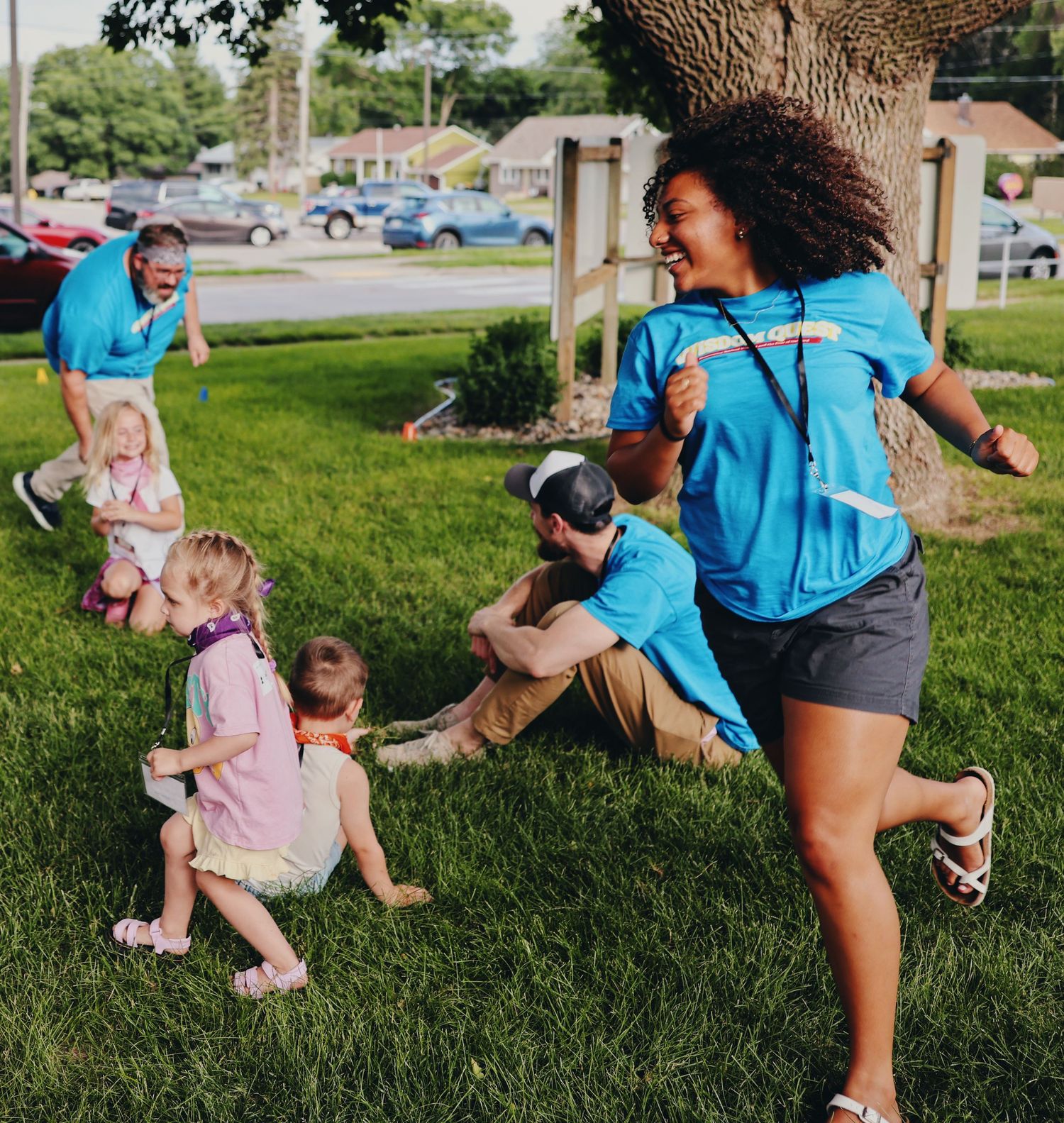 Adults and children playing outdoors on green grass near a tree, wearing matching blue shirts on a sunny day.