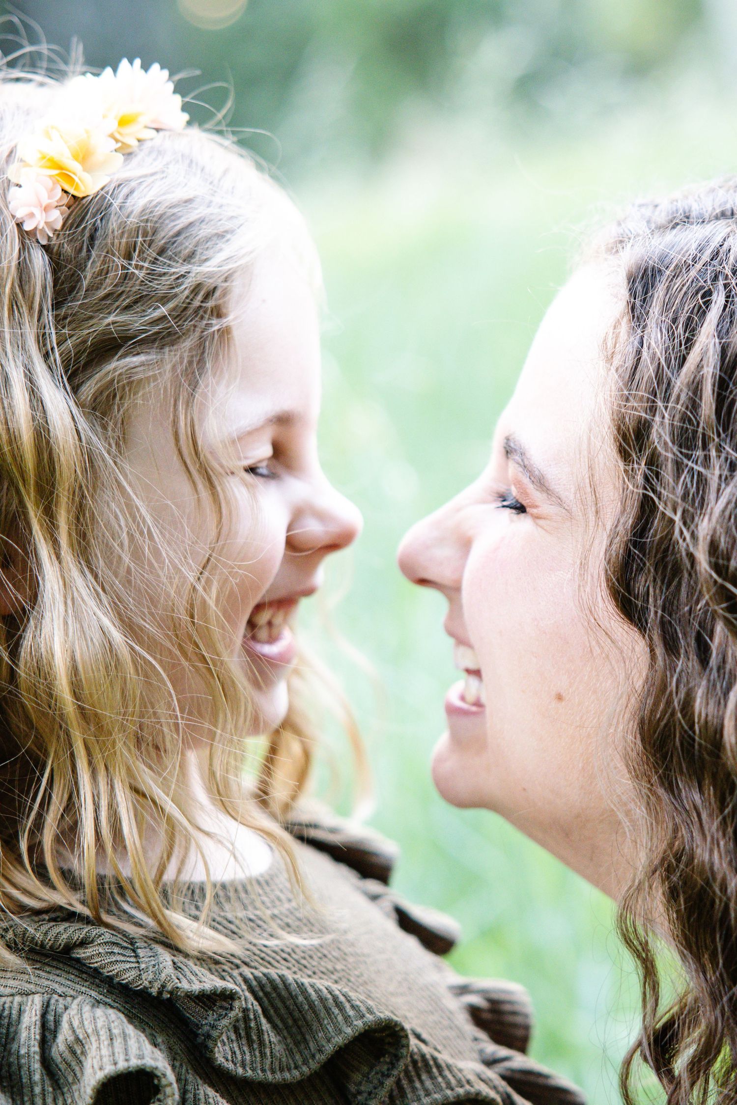A young girl with flower hair accessories smiles face-to-face with a curly-haired woman outdoors.