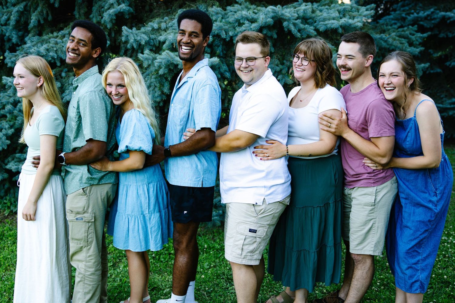 A diverse group of eight young adults laughing and posing in a line outdoors near a large evergreen tree.