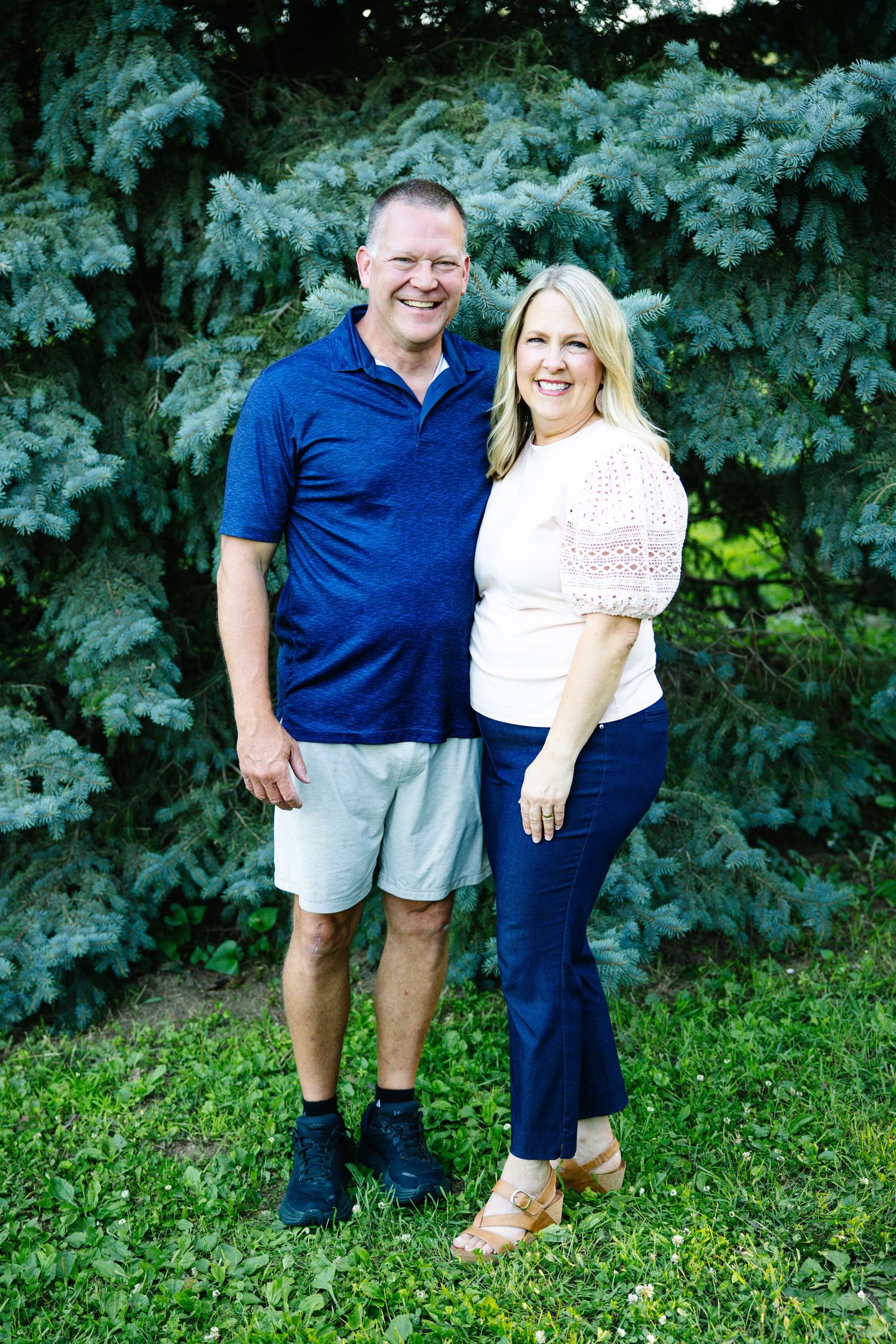 Smiling couple posing together outdoors in front of lush blue spruce trees on a sunny day.