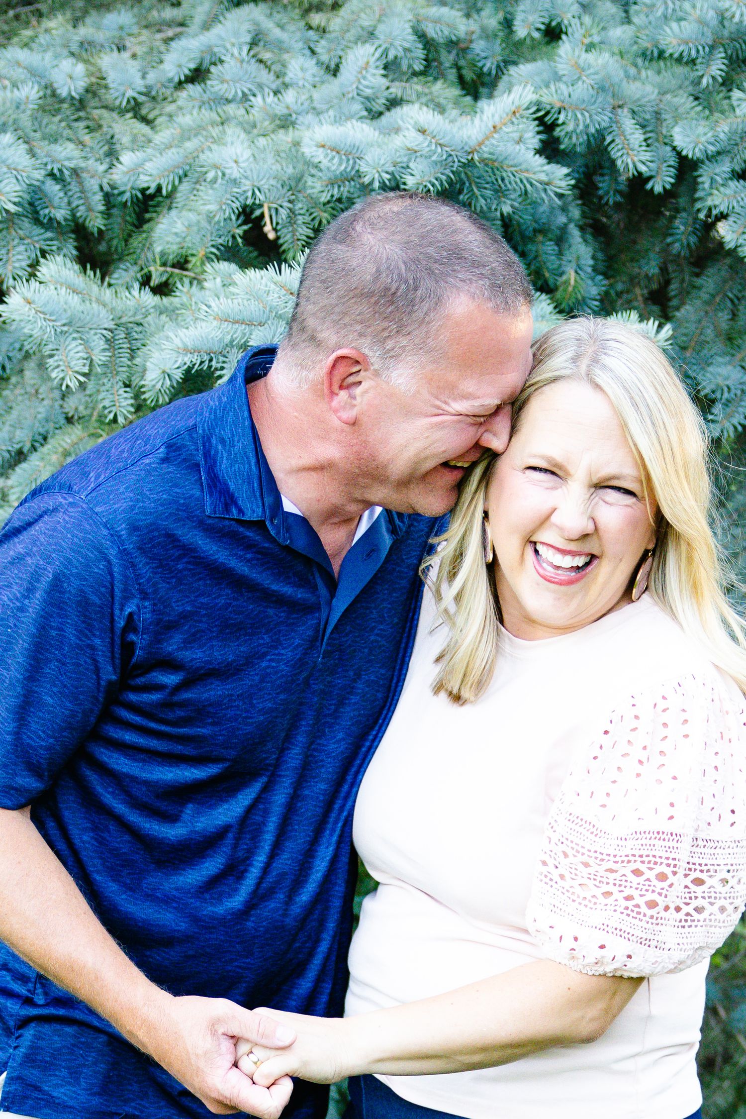 A laughing couple holds hands outdoors near a blue spruce tree, sharing a joyful and affectionate moment together.
