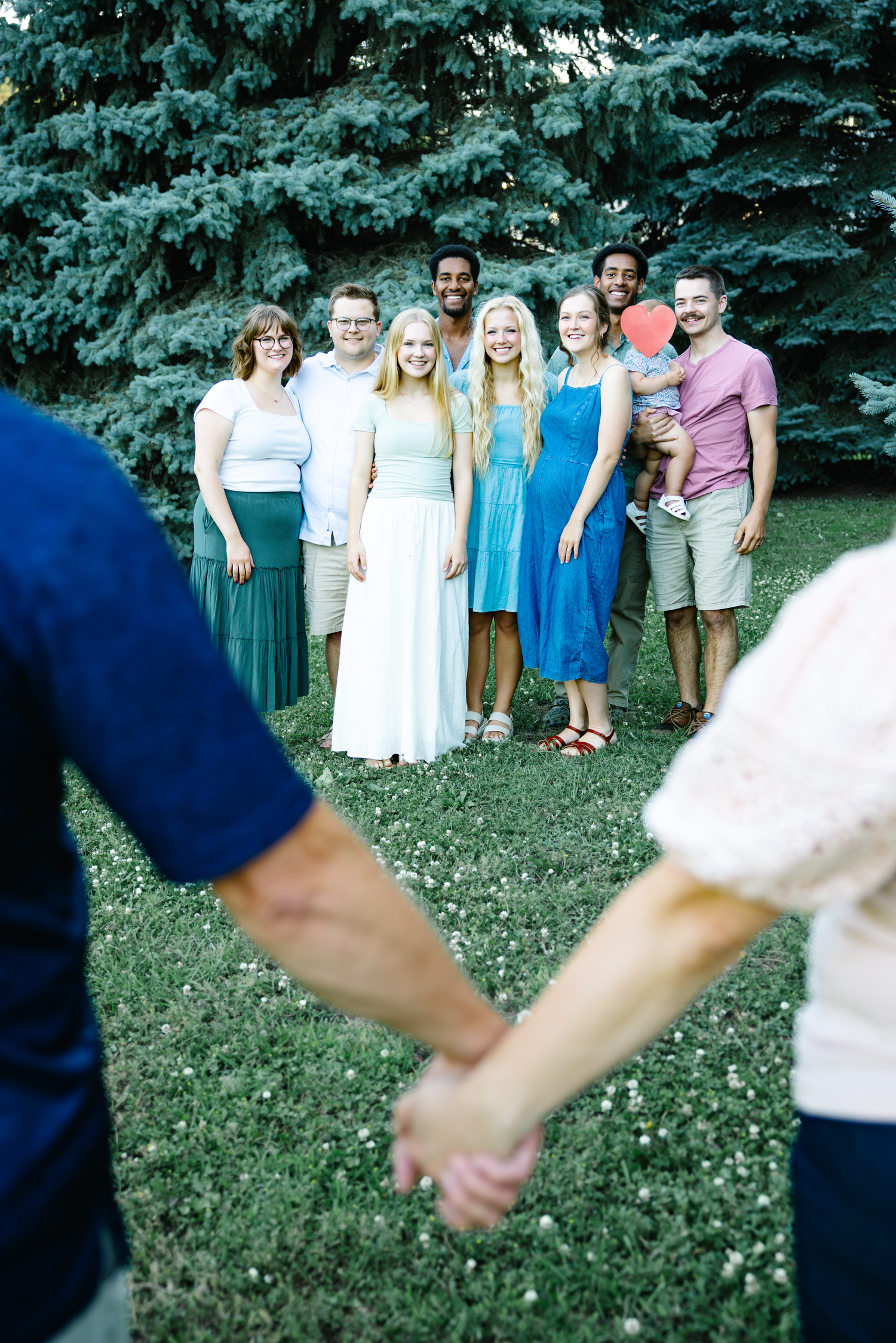 A couple holds hands in the foreground while a group of friends poses on a green lawn with evergreen trees behind them.