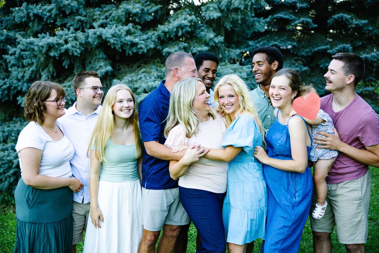 Large family group laughing and embracing outdoors near evergreen trees on a sunny summer day.