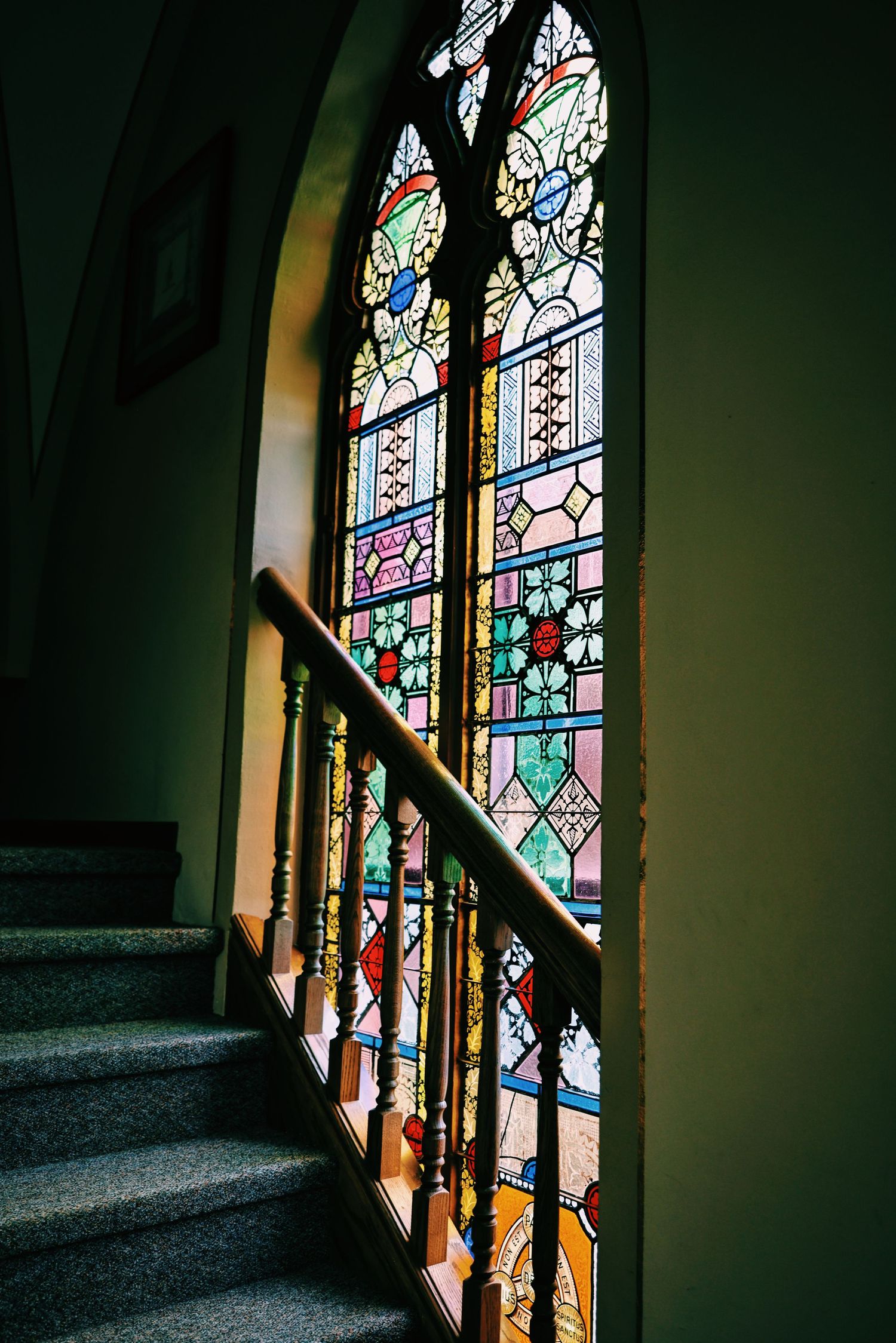 Colorful stained glass Gothic window illuminating a wooden staircase railing in an elegant historic building interior.